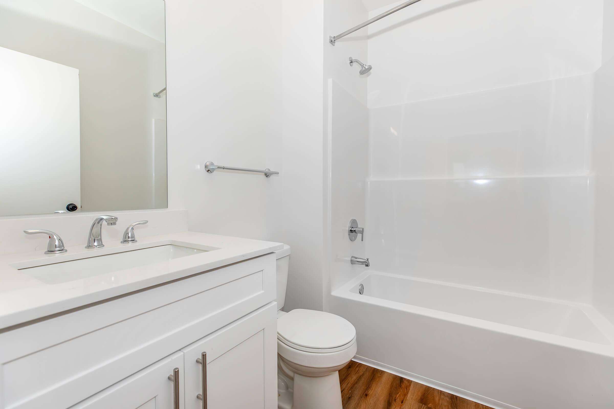 A modern bathroom featuring a white bathtub, a sleek vanity with a mirror, and a toilet. The walls are painted white, and the floor is made of laminate wood. Stainless steel fixtures are visible, providing a clean and contemporary look.