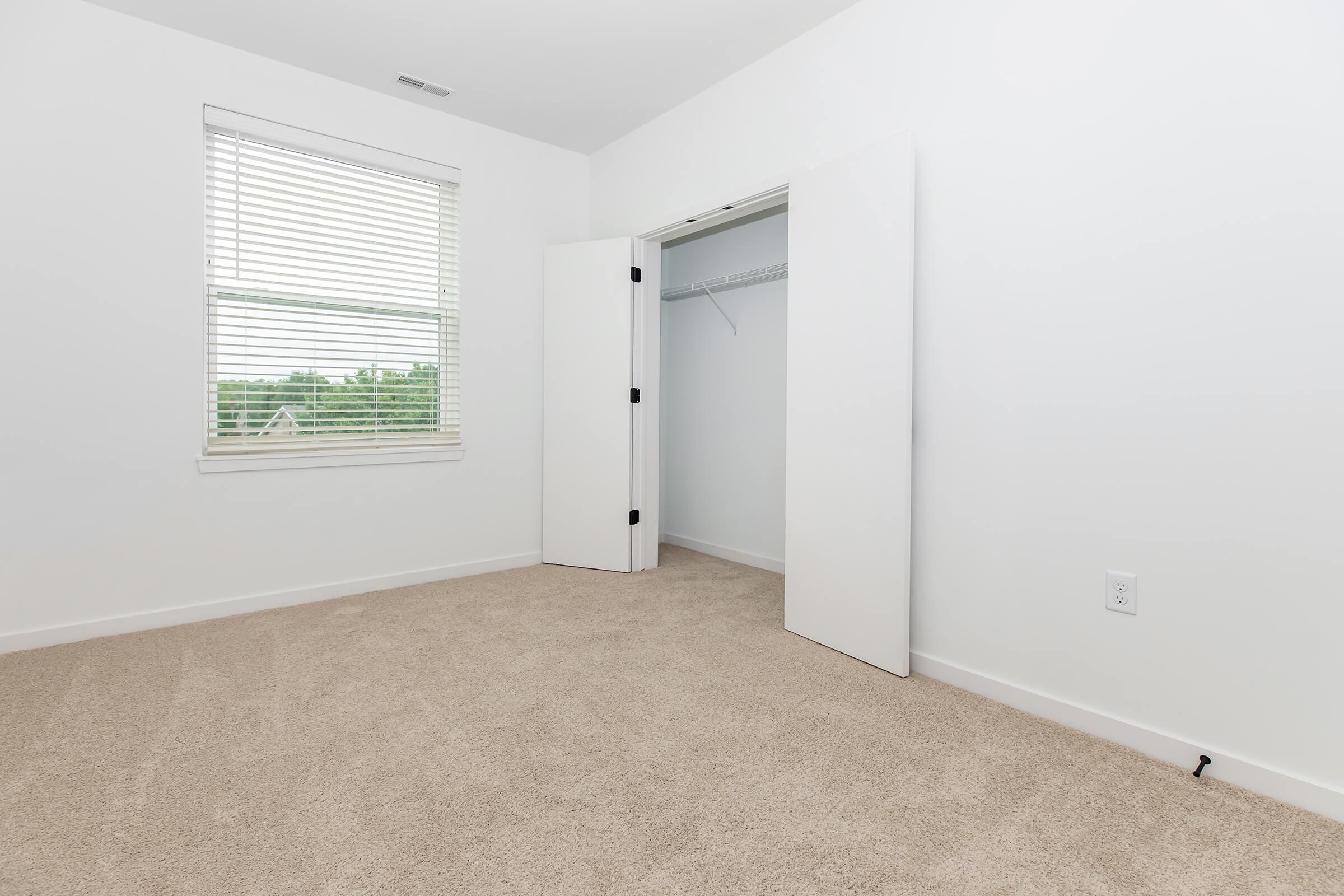 A clean, empty bedroom with light beige carpet and white walls. There is a window with blinds allowing natural light to enter. The closet features double doors that are open, revealing a simple interior with a hanging rod. The room is minimally furnished, showcasing a spacious and airy atmosphere.