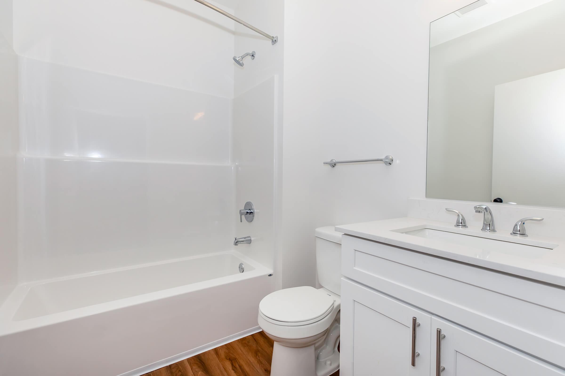 A modern bathroom featuring a white tub and shower combination, a white vanity with a sink and three faucets, a white toilet, and a large mirror. The walls are painted white and the floor is made of wooden planks.