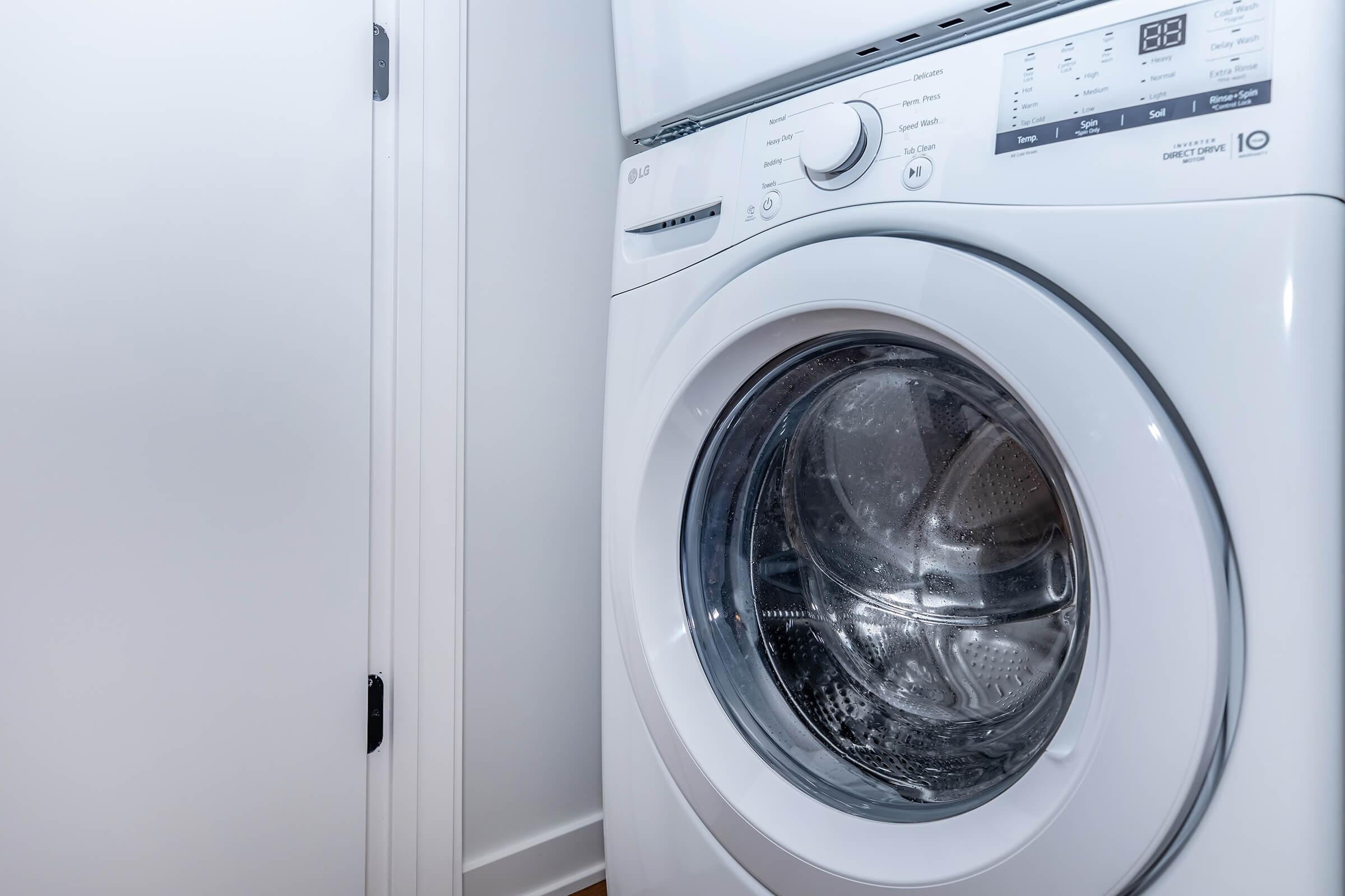 Close-up of a modern white washing machine with a transparent front door, showcasing its drum inside. The machine is positioned against a wall, with a door nearby, emphasizing a clean and organized laundry space. The control panel displays various settings.