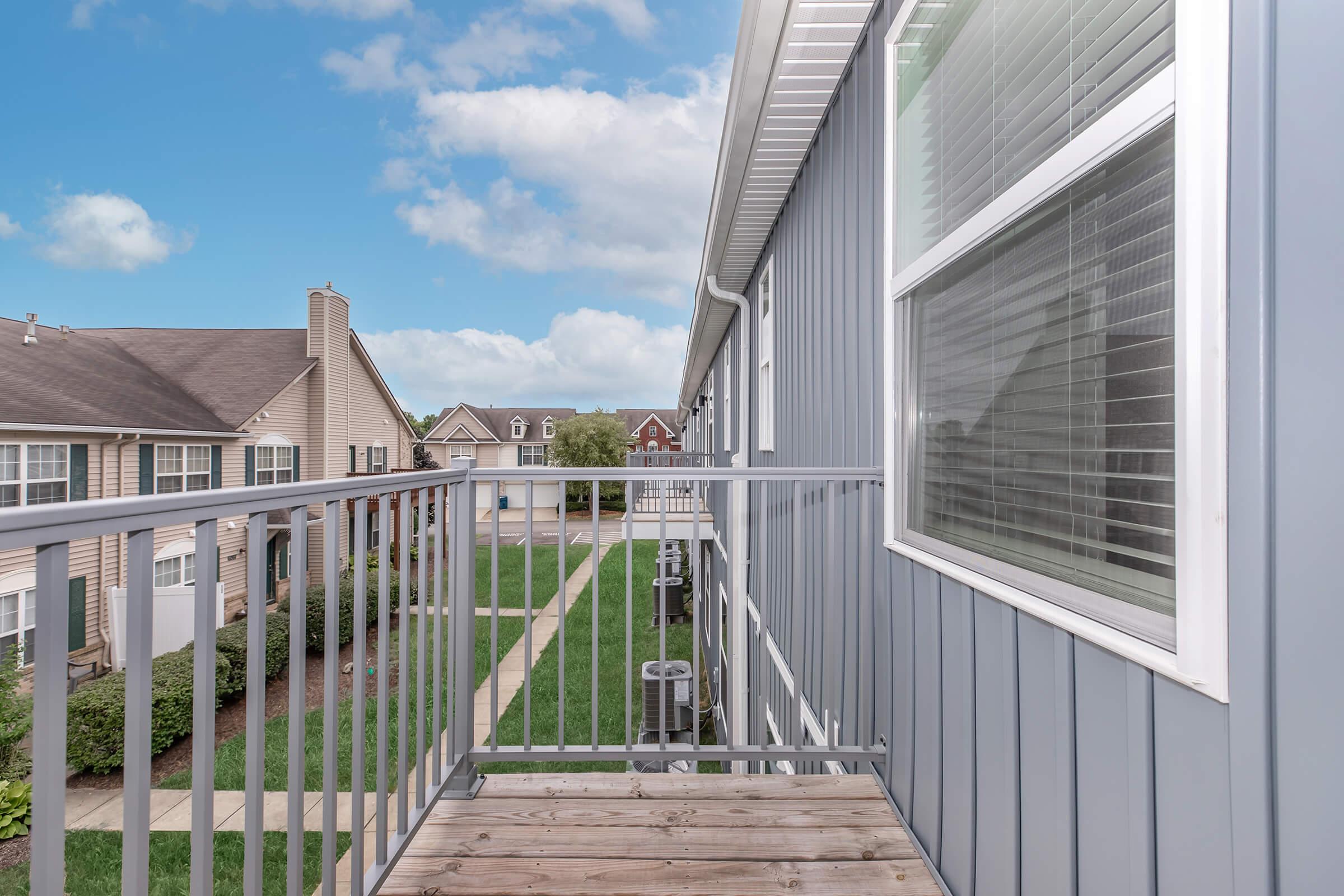 View from a balcony overlooking a landscaped courtyard and neighboring buildings. The scene features a gray exterior house wall, a metal railing, and a clear blue sky with scattered clouds. Pathways and green grass are visible below, providing a serene residential atmosphere.