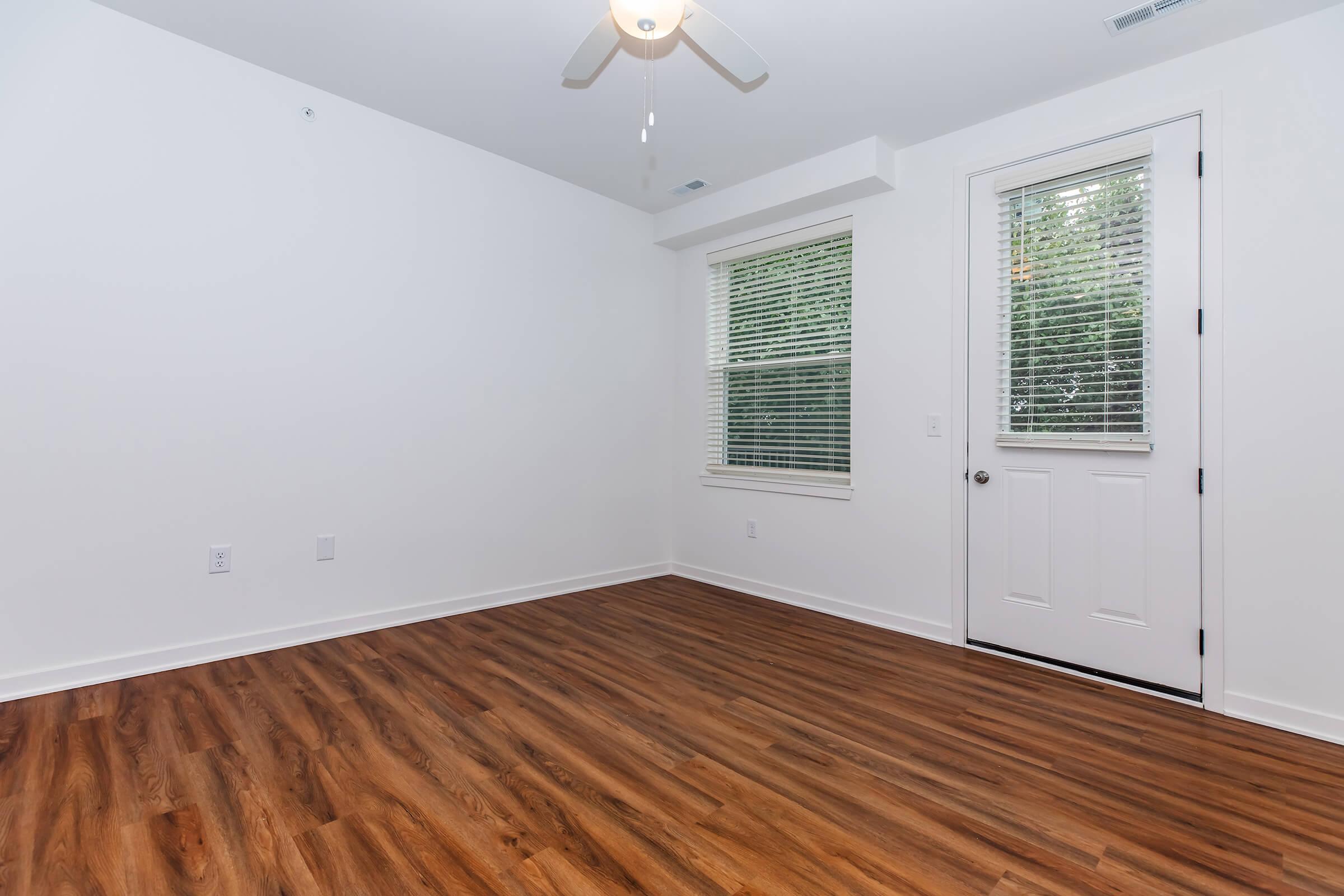 Empty room with hardwood flooring, a ceiling fan, and a single window. There is a white door leading outside, and the walls are painted in a light color. Natural light is coming through the window, creating a bright atmosphere.