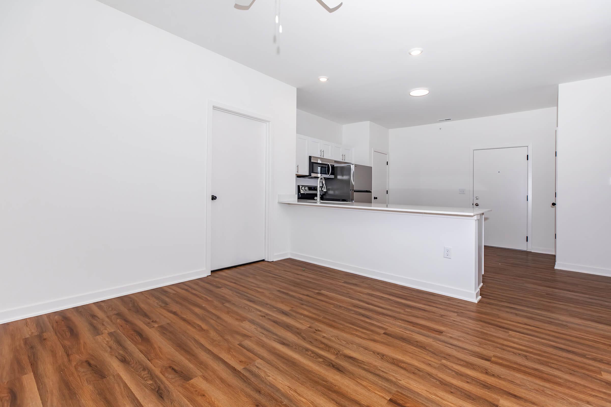 An empty apartment interior featuring a kitchen area with a counter and modern appliances, light-colored walls, a ceiling fan, and wooden flooring. The space is well-lit with overhead lights, showcasing an open layout that connects the kitchen to the living area.