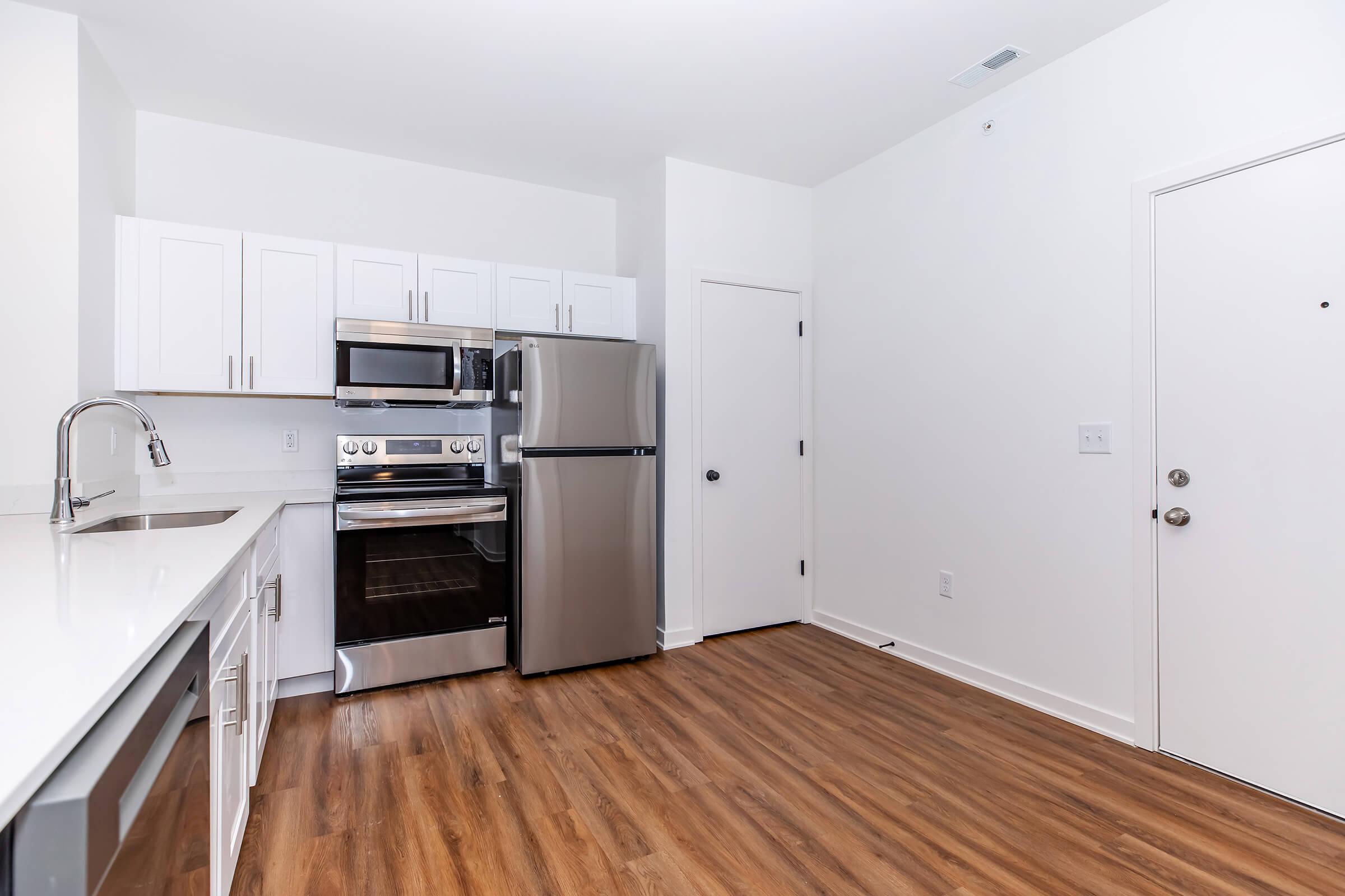 Modern kitchen with stainless steel appliances including a microwave, oven, and refrigerator, set against white cabinets. The space features a light wood floor and a door leading to another room, along with an entrance door. The walls are painted white, creating a bright and clean atmosphere.