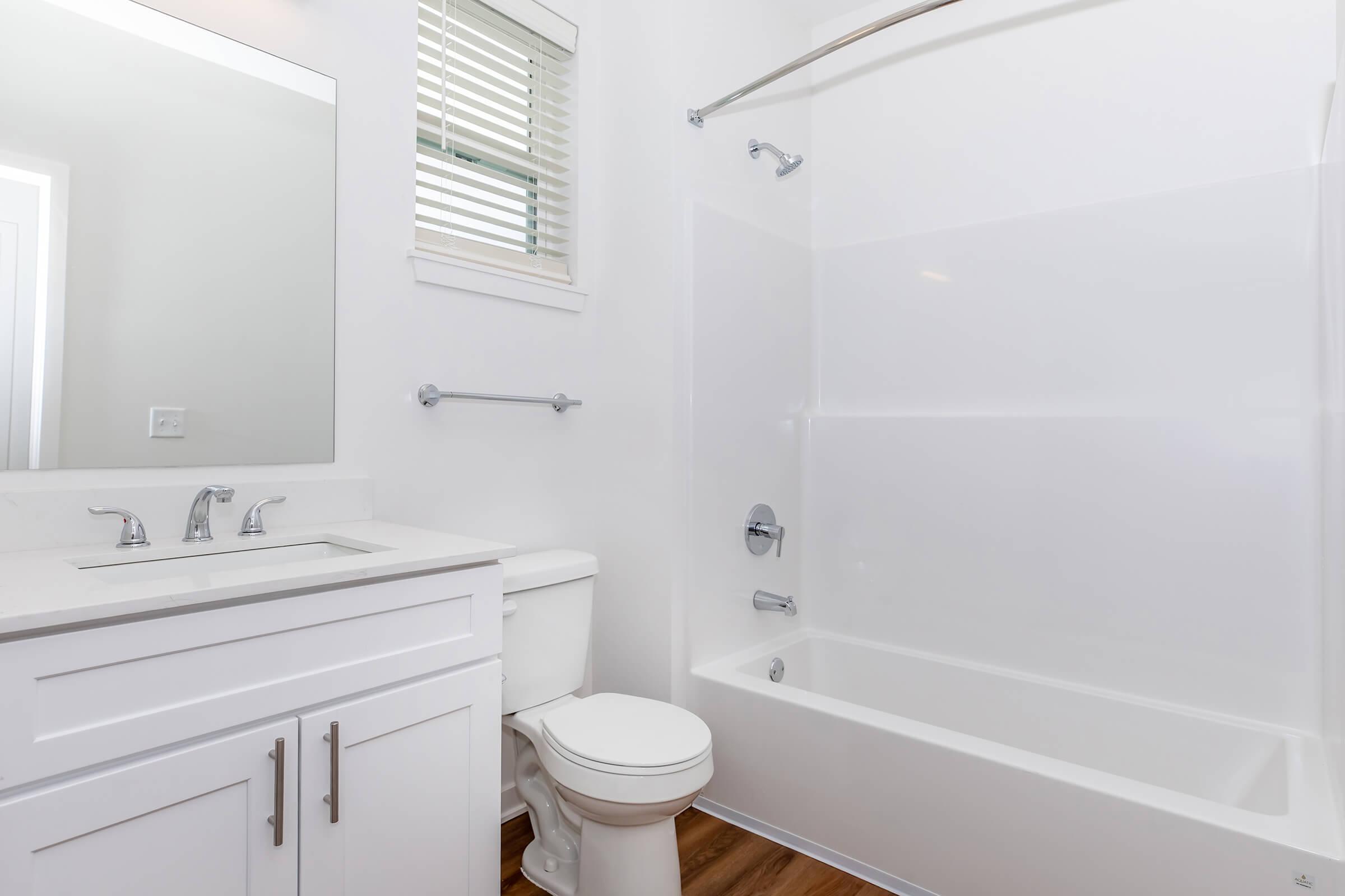 A bright, modern bathroom featuring a white bathtub with a shower, a toilet, and a sink with a large mirror above it. The walls are painted white, and there’s natural light coming through a small window with blinds. The flooring is wood, adding warmth to the space.