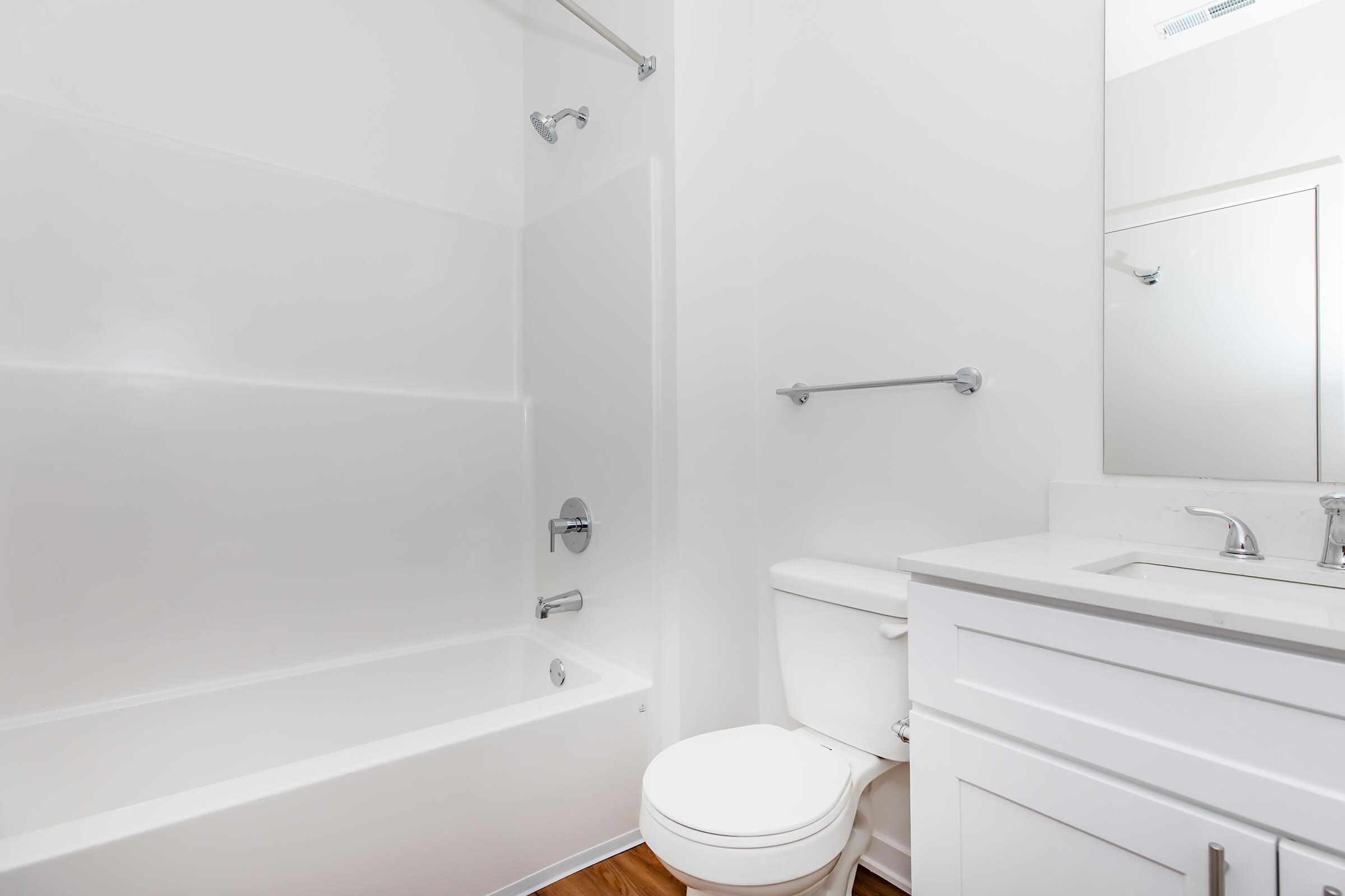 A clean, modern bathroom featuring a white bathtub with a shower, a toilet, and a sink with a mirror above it. The walls are painted white, and the floor is made of light-colored wood. Simple, functional design with minimal decor.