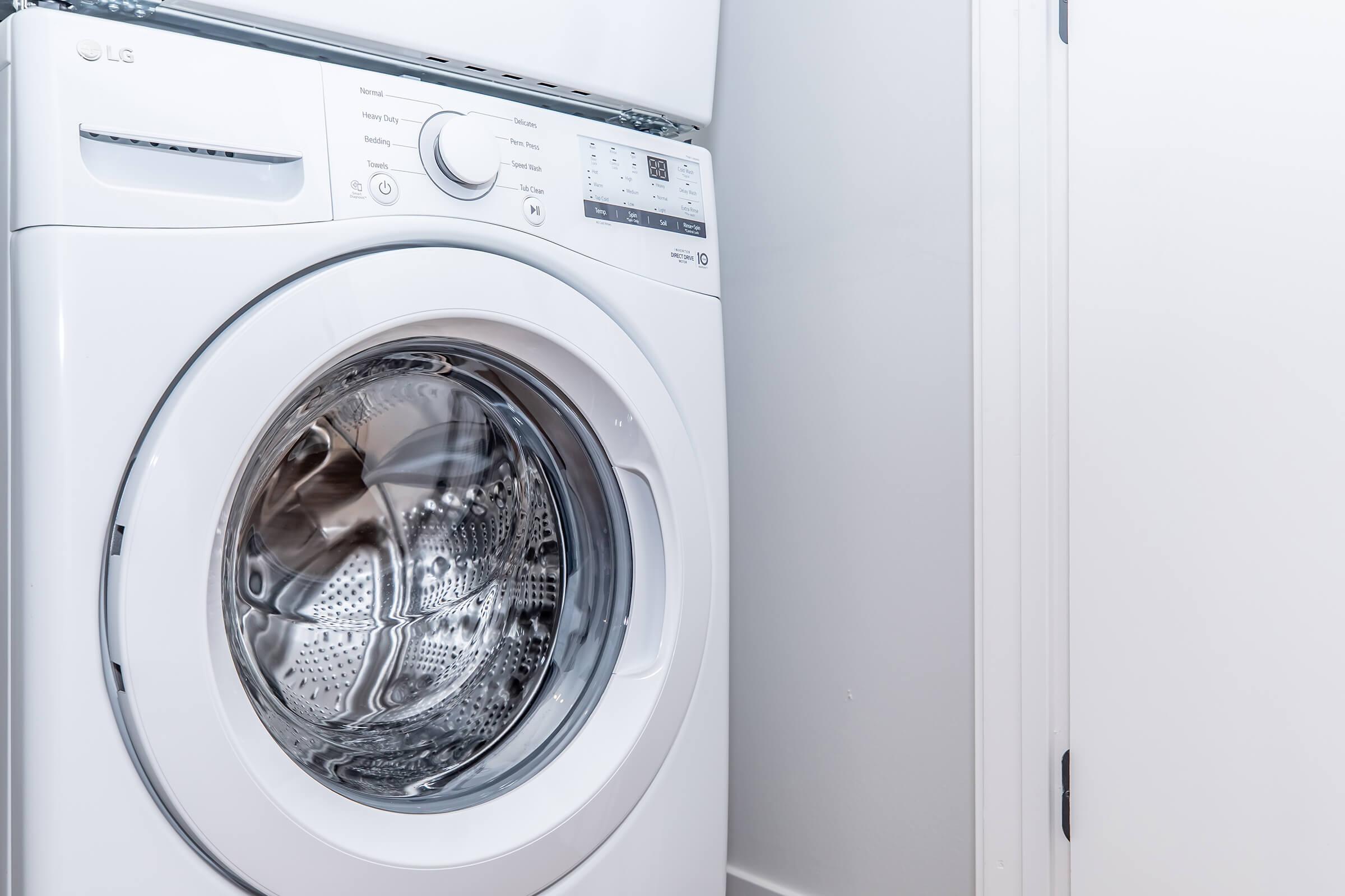 A close-up view of a white front-loading washing machine, partially visible in a laundry room setting. The machine features a circular glass door, control panel with various buttons and settings, and is positioned next to a wall with a door.