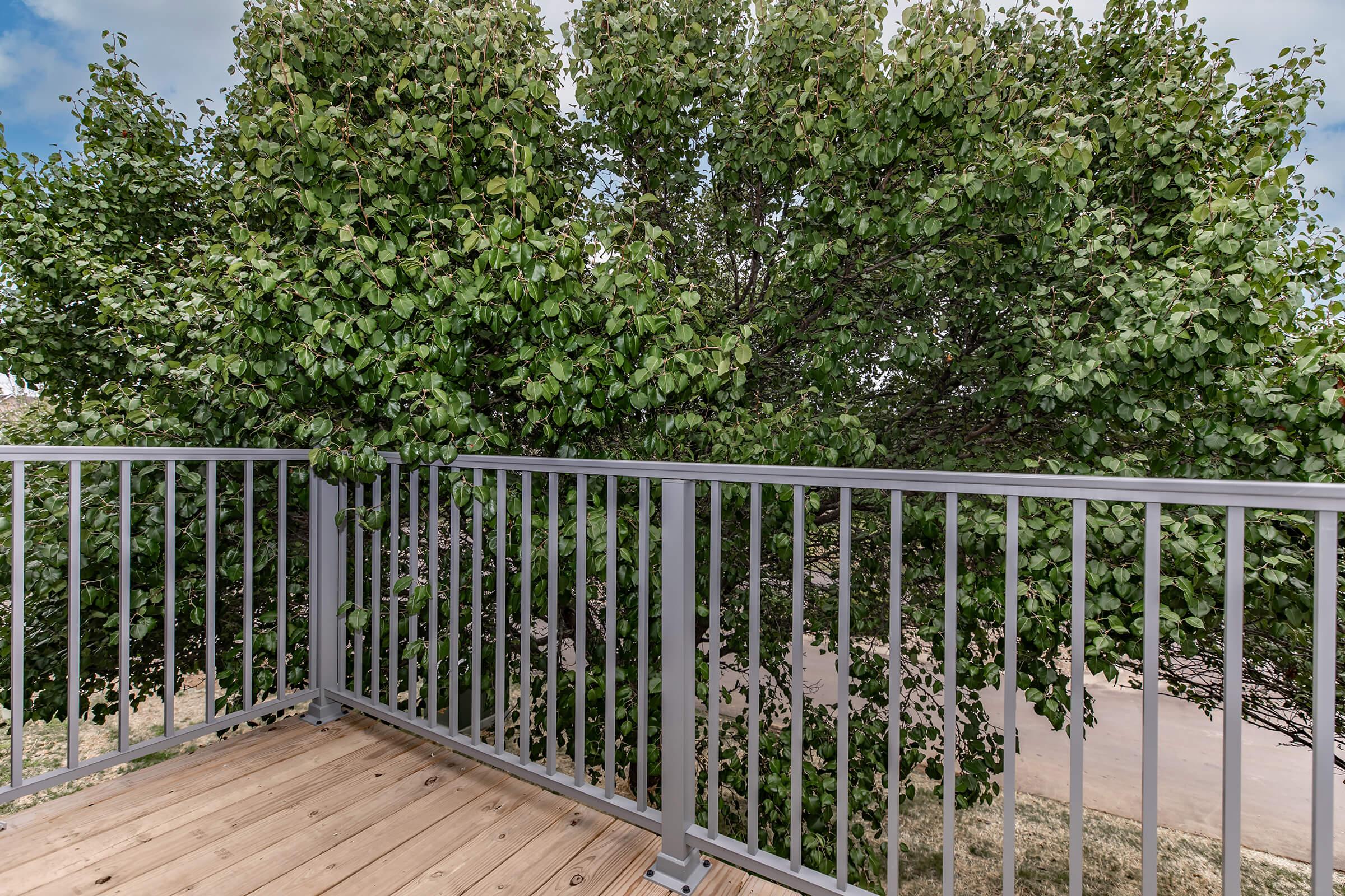 A view from a balcony featuring a wooden deck and a metal railing, with a large leafy tree partially obstructing the view. The background shows a cloudy sky and hints of a landscape beyond the tree.