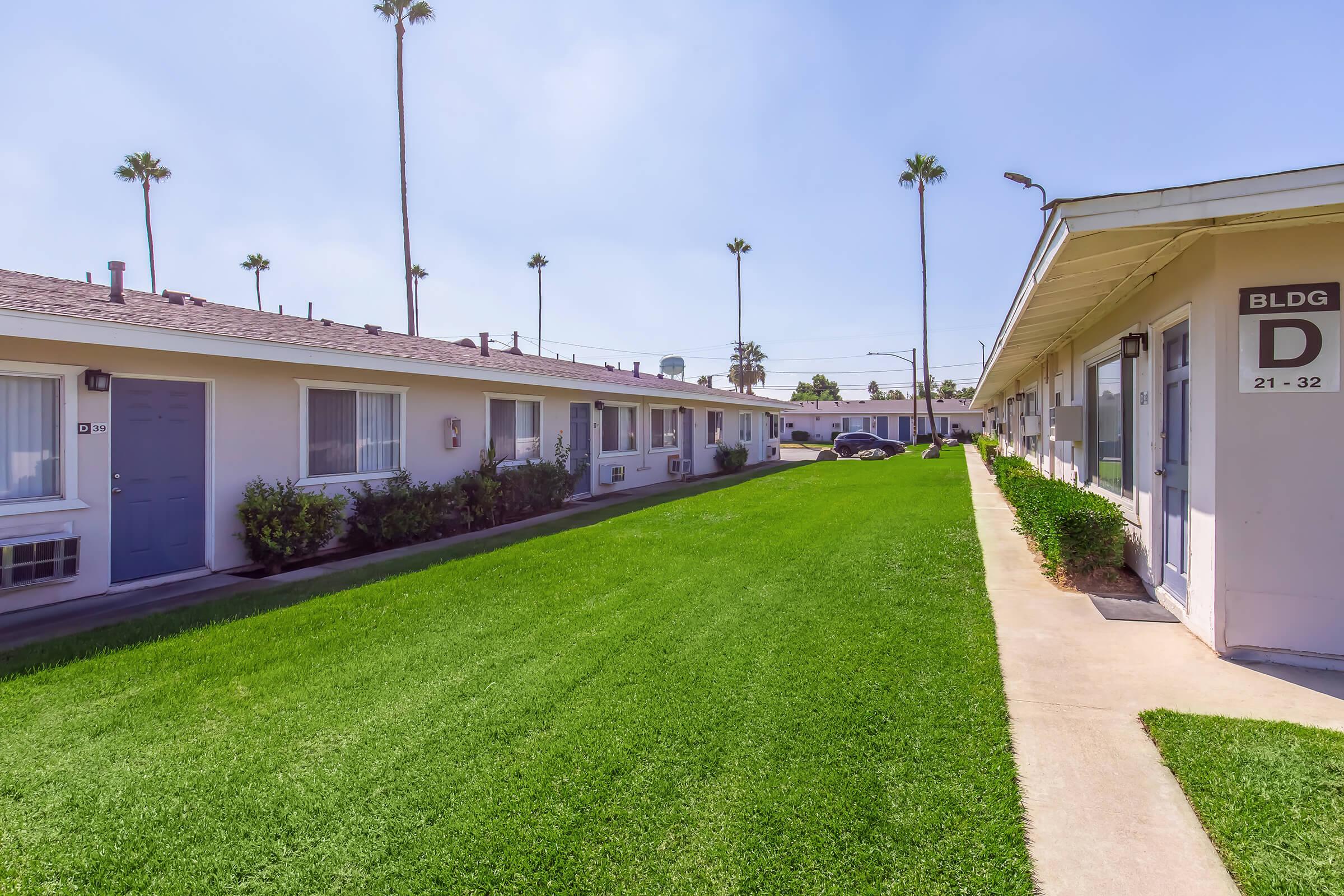View of a landscaped courtyard in a multi-unit apartment complex, featuring green grass and two rows of single-story buildings. Palm trees line the background under a clear blue sky. The scene conveys a sunny and inviting residential atmosphere.