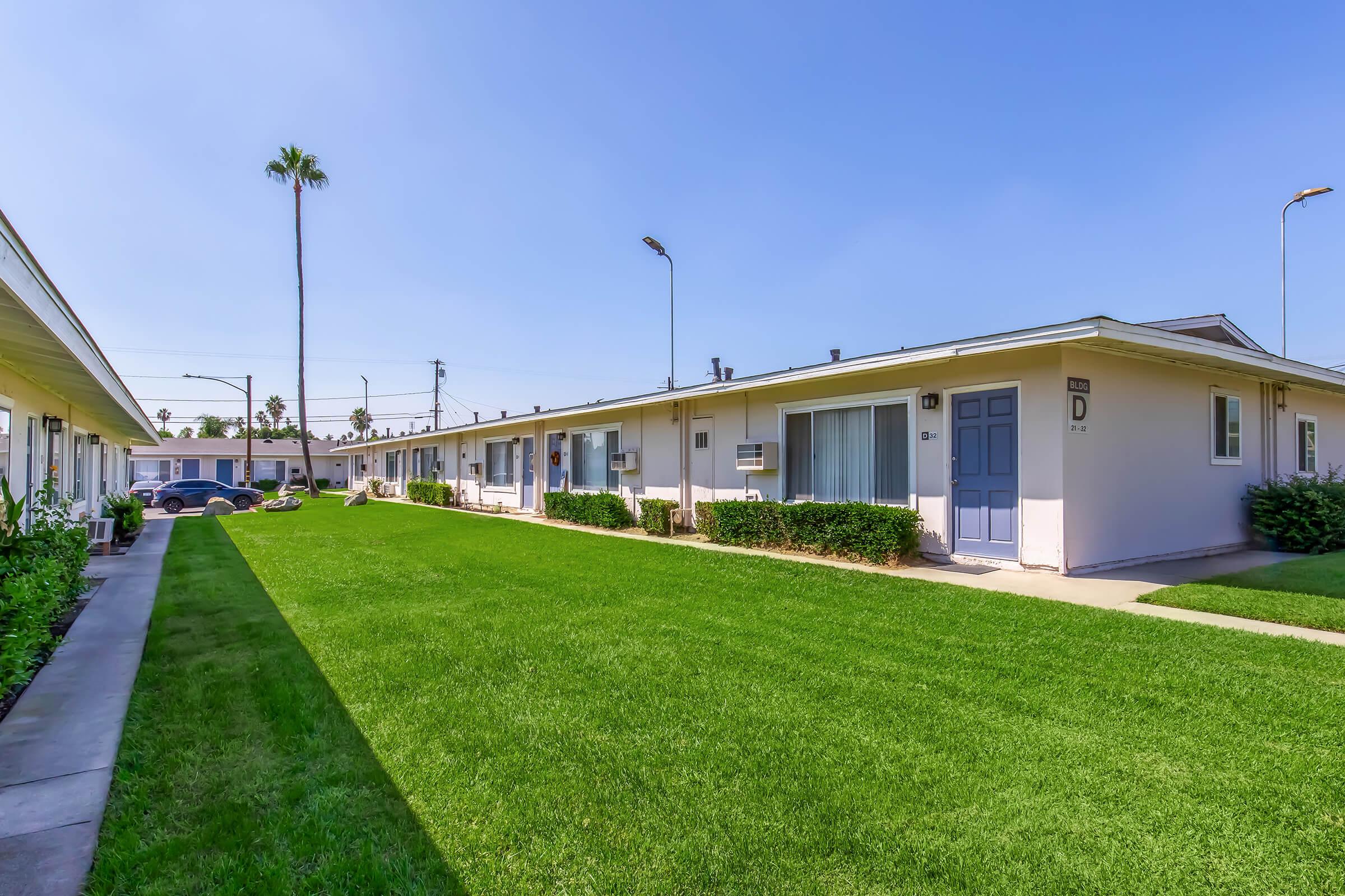 A view of a single-story apartment complex with a well-maintained grassy area in front. Several units are visible, featuring pastel-colored walls and blue doors. Palm trees line the background under a clear blue sky. The pathway is bordered by greenery, creating a serene residential atmosphere.