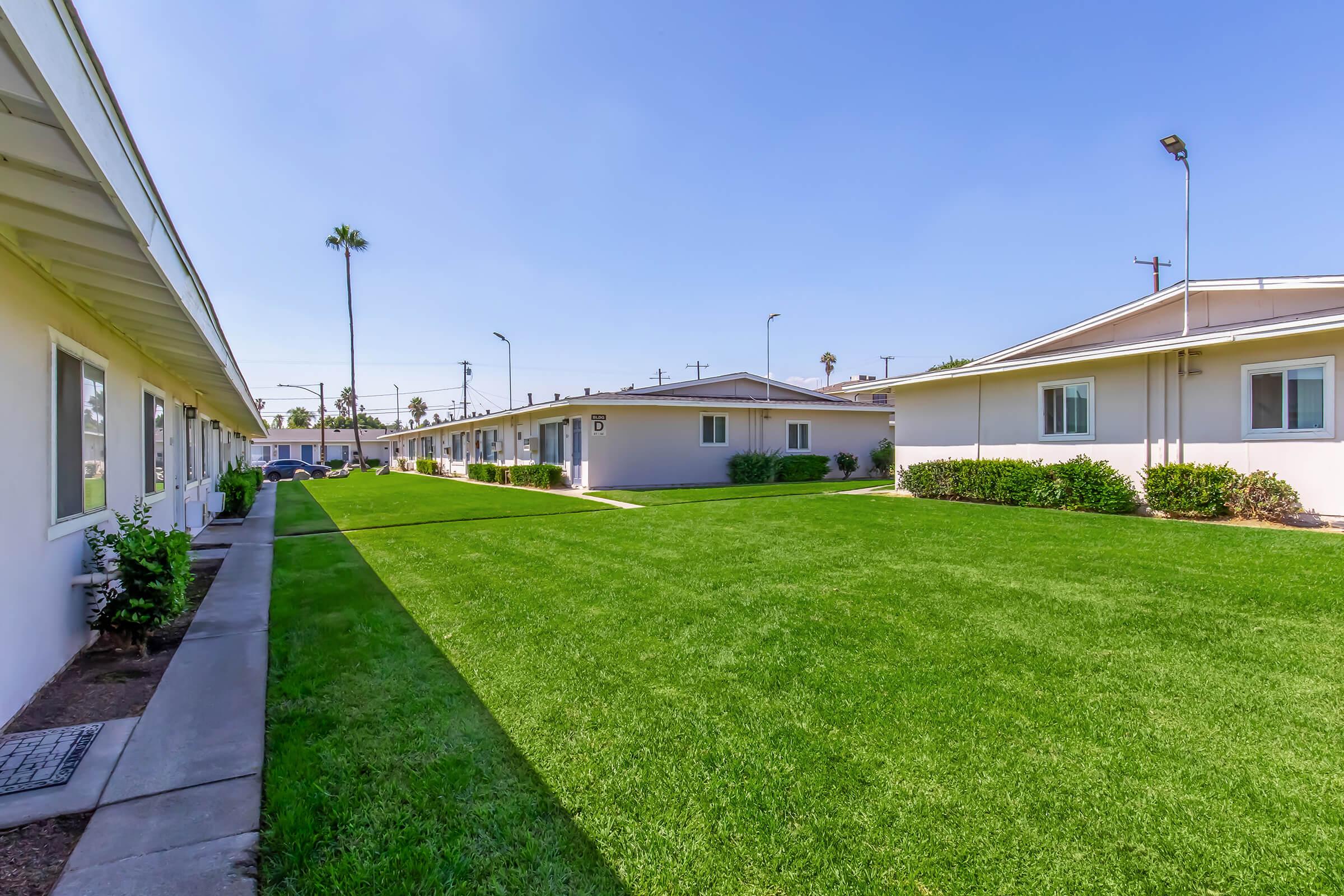 View of a well-maintained grassy area between two rows of buildings. The scene features palm trees in the background and clear blue skies. The buildings are low-rise with windows and are situated in a neat, open layout, providing a spacious and inviting atmosphere.