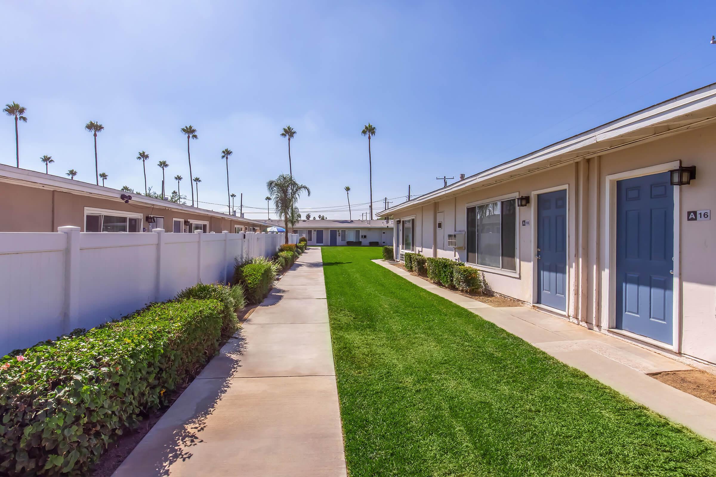 A well-maintained pathway lined with green grass and neat hedges, leading between two rows of single-story buildings. Each building features a doorway with blue doors, and palm trees are visible against a clear blue sky. The scene conveys a peaceful and inviting atmosphere.