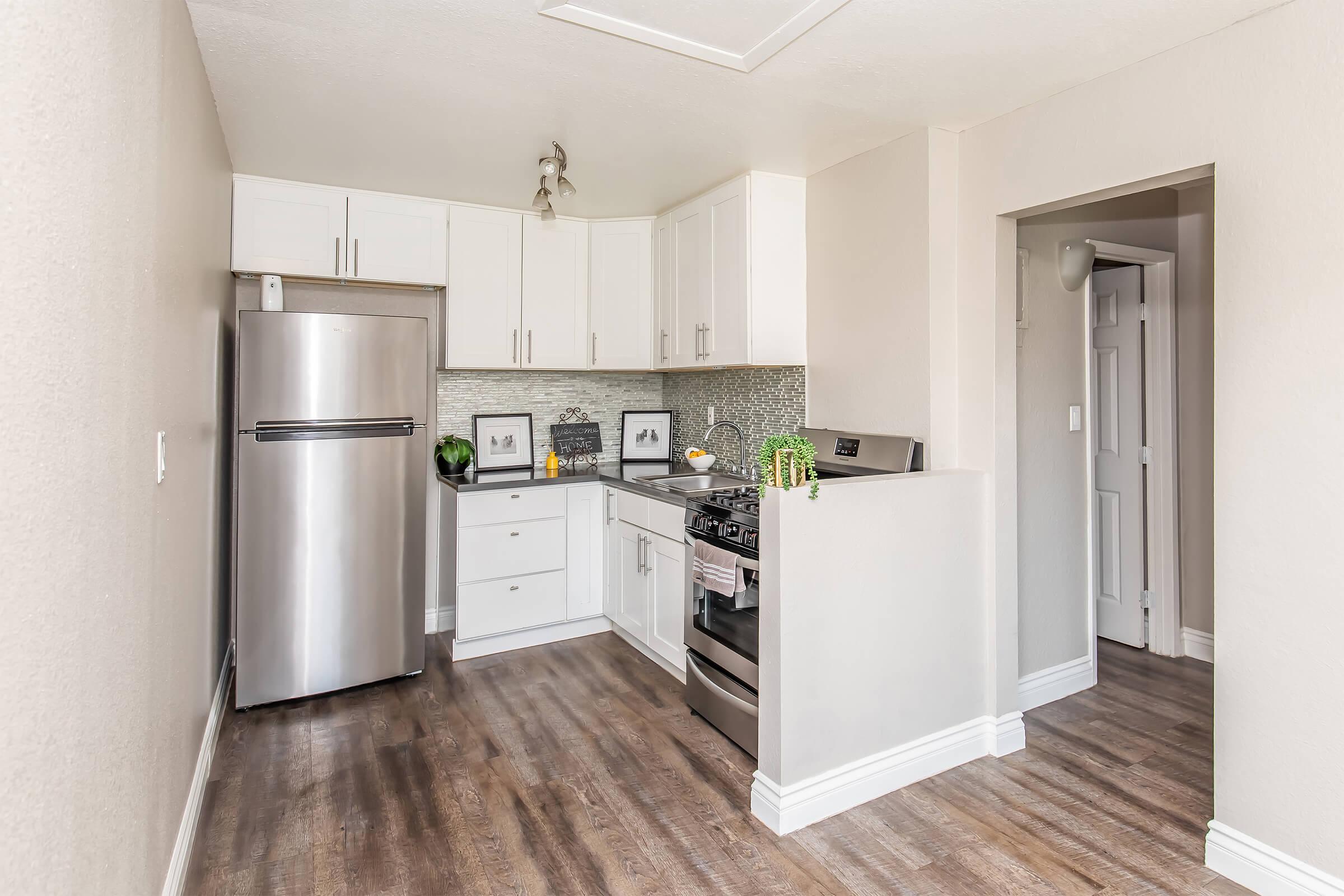 A modern kitchen featuring stainless steel appliances, including a refrigerator and a stove. White cabinets with a sleek countertop are complemented by a stylish backsplash. The space has wooden flooring and bright lighting, creating a clean and inviting atmosphere.