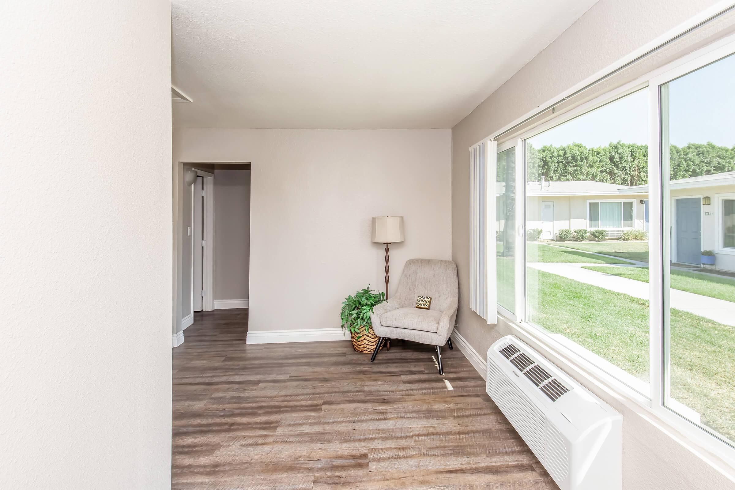 A well-lit interior space featuring a beige wall, wooden flooring, and a cozy armchair beside a lamp. A large window offers a view of grassy outdoor area and nearby buildings. A small potted plant adds a touch of greenery to the room.