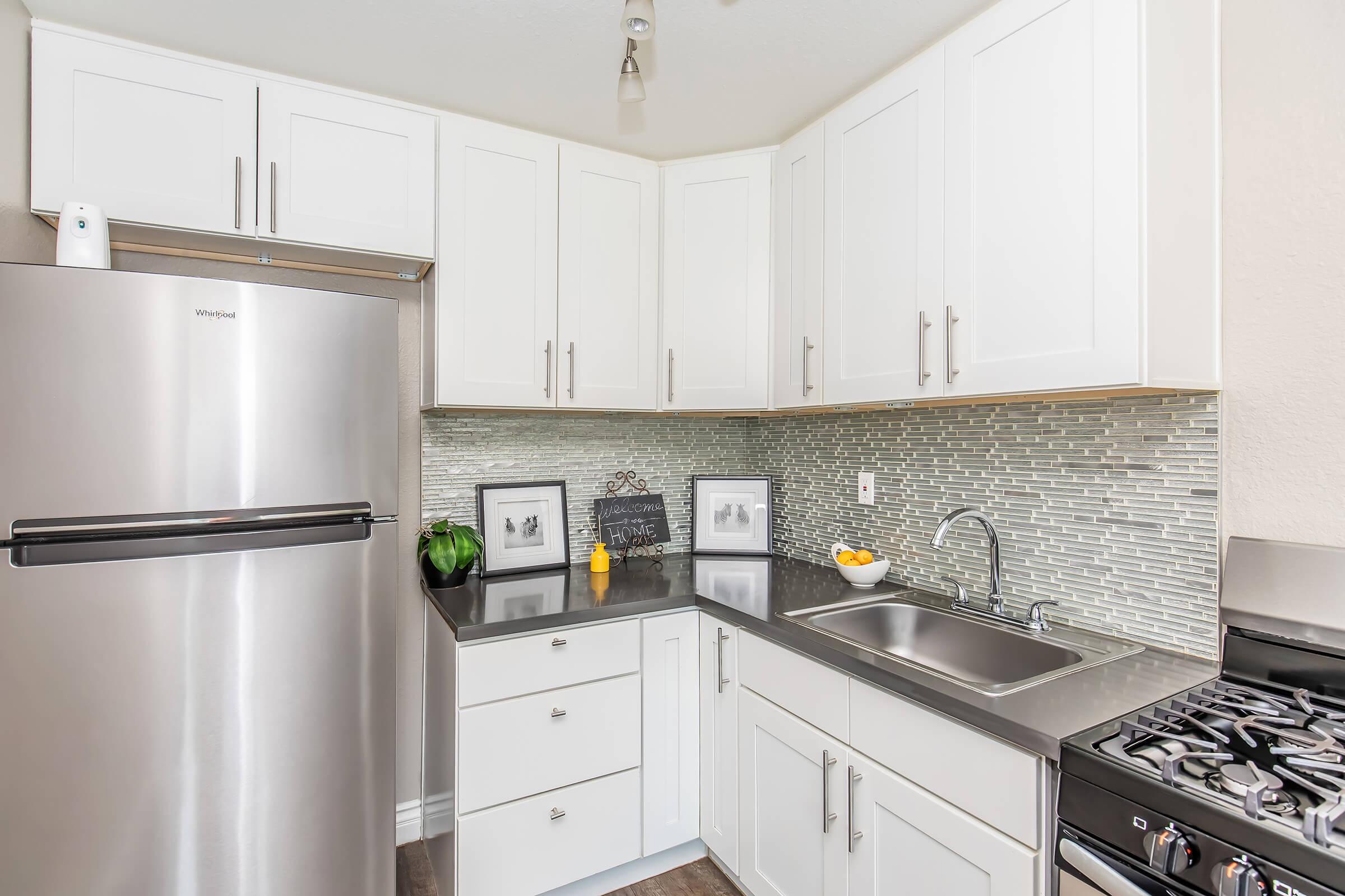 A modern kitchen featuring white cabinetry, a stainless steel refrigerator, and a sink with a chrome faucet. The countertop is dark and there are decorative elements, including framed pictures and a small plant. The backsplash consists of small, glass tiles, and there is a gas stove next to the sink.