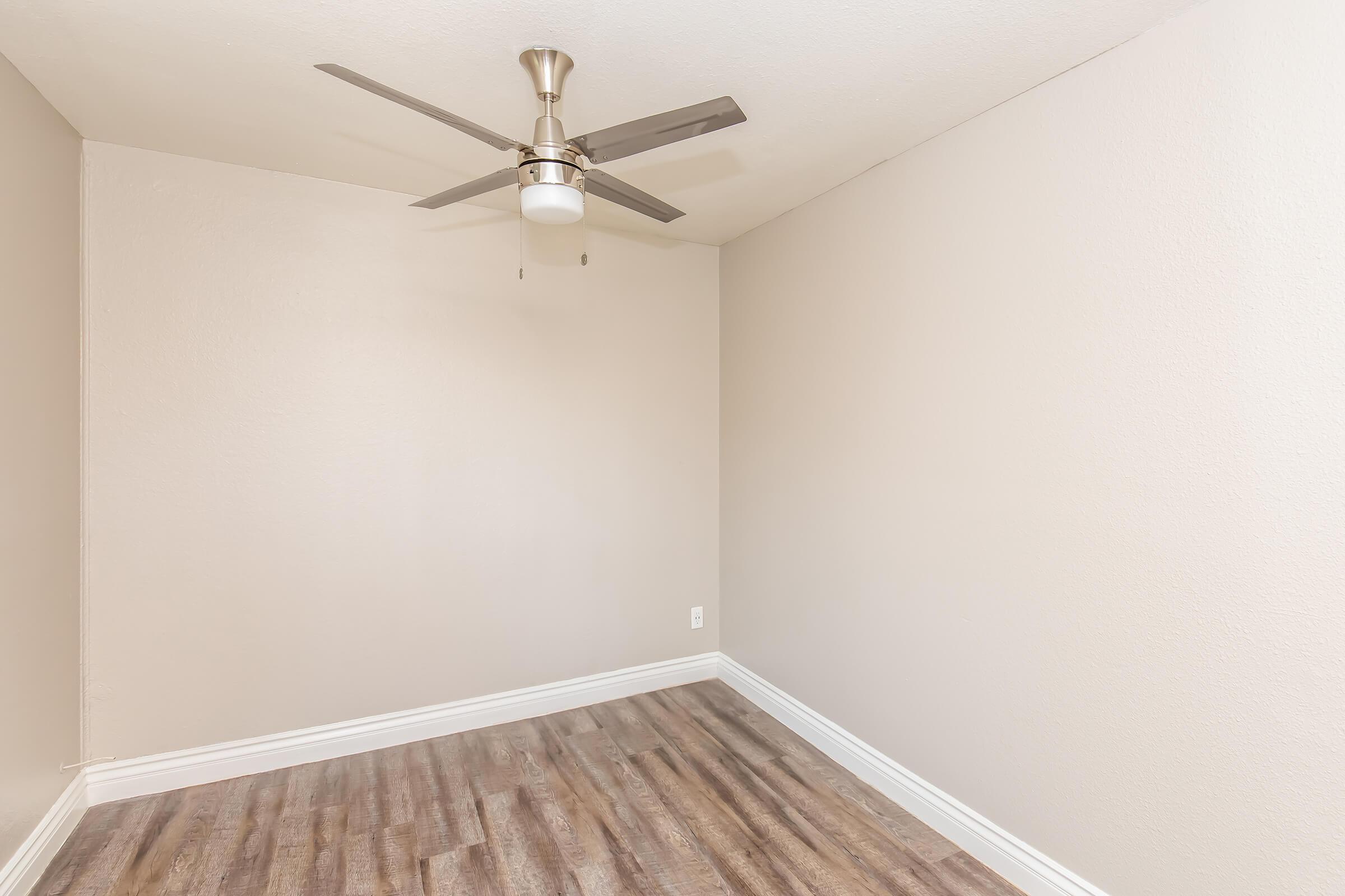 Empty room with light brown walls and a light wood floor. A ceiling fan with a light fixture is mounted in the center of the ceiling. The room has a simple, open layout suitable for various uses or furnishings.