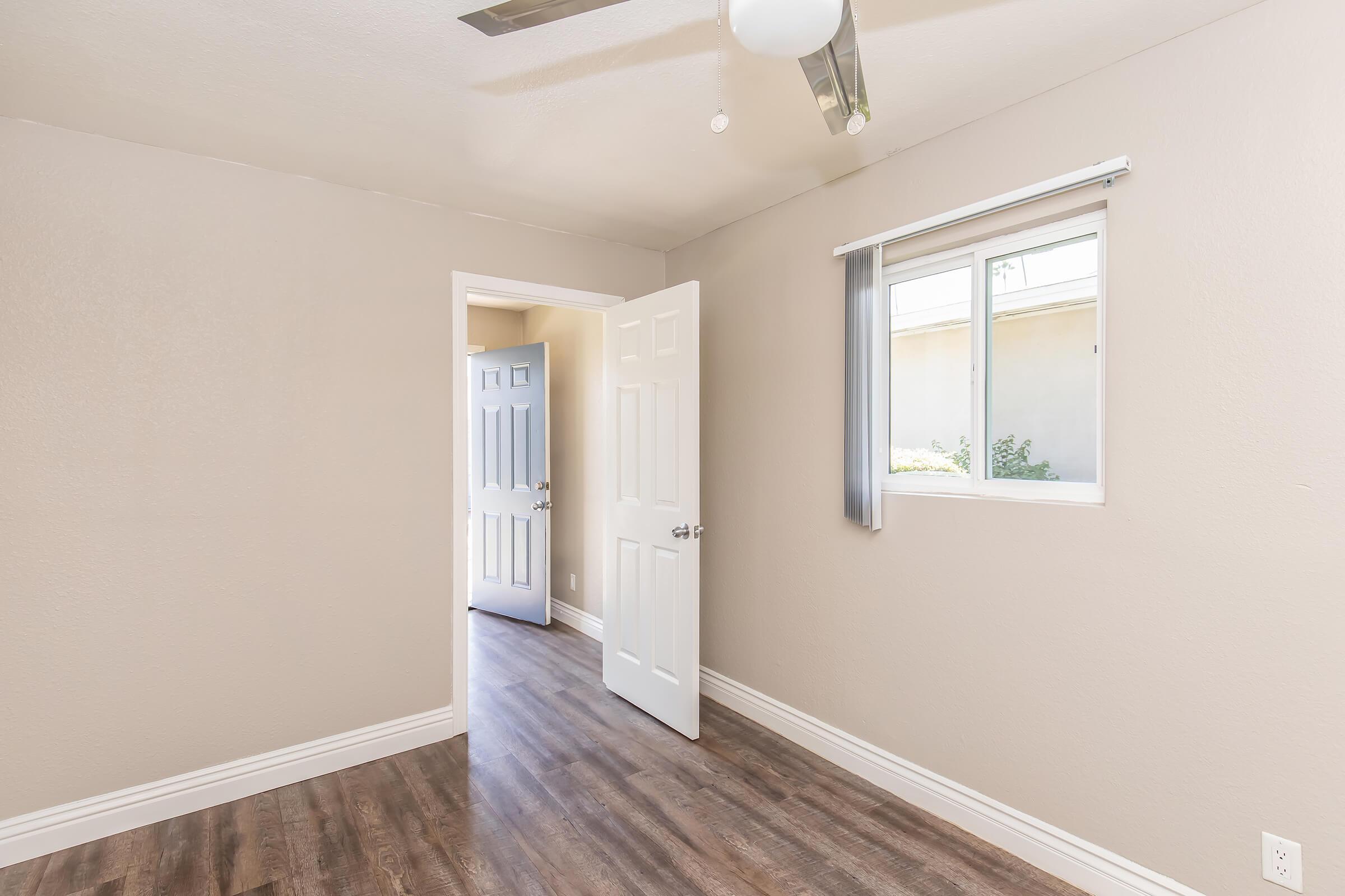 A well-lit, empty room featuring light gray walls and a ceiling fan. A door on the right leads to another room, while a window allows natural light to enter. The floor is covered with hardwood laminate, enhancing the spacious feel of the area.
