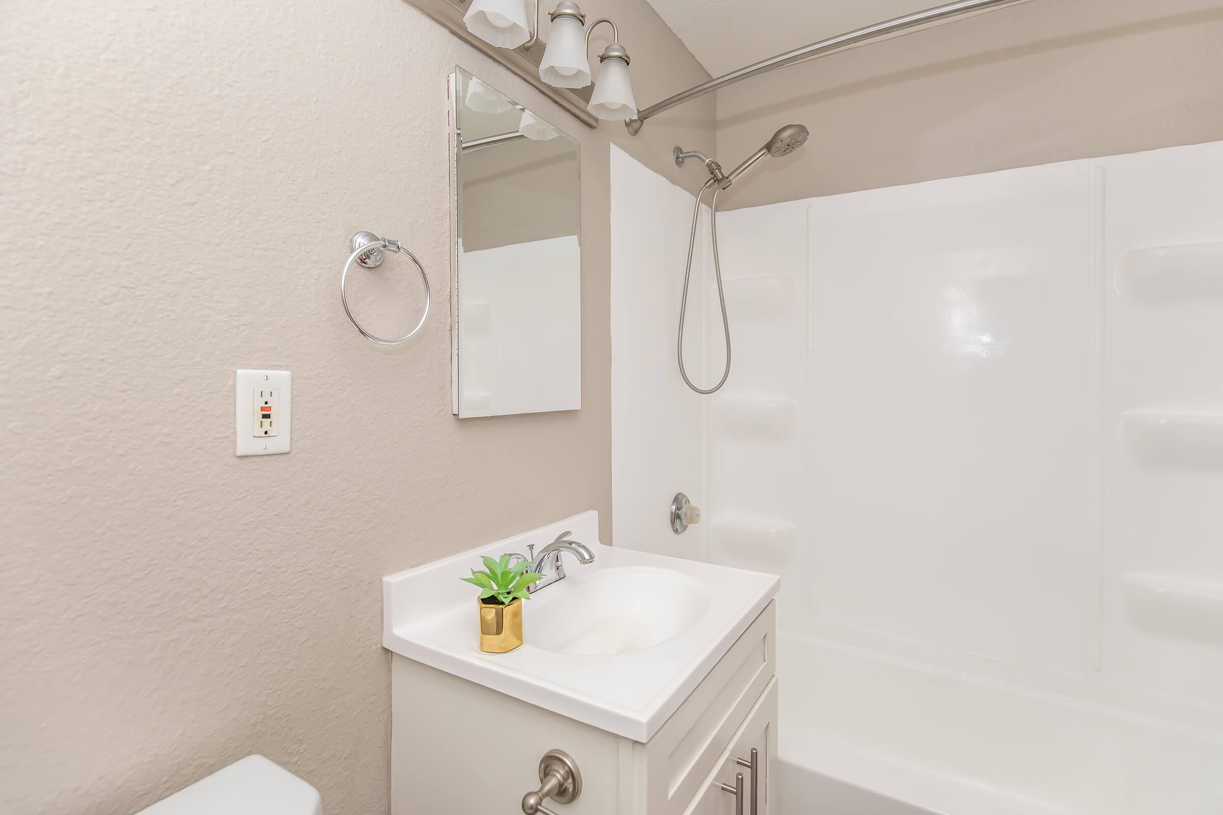 A clean and modern bathroom featuring a white shower/tub unit, a small white sink with a gold plant pot on the countertop, a wall-mounted mirror, and light fixtures. The walls are painted in a neutral tone, and a towel ring is visible beside the sink.