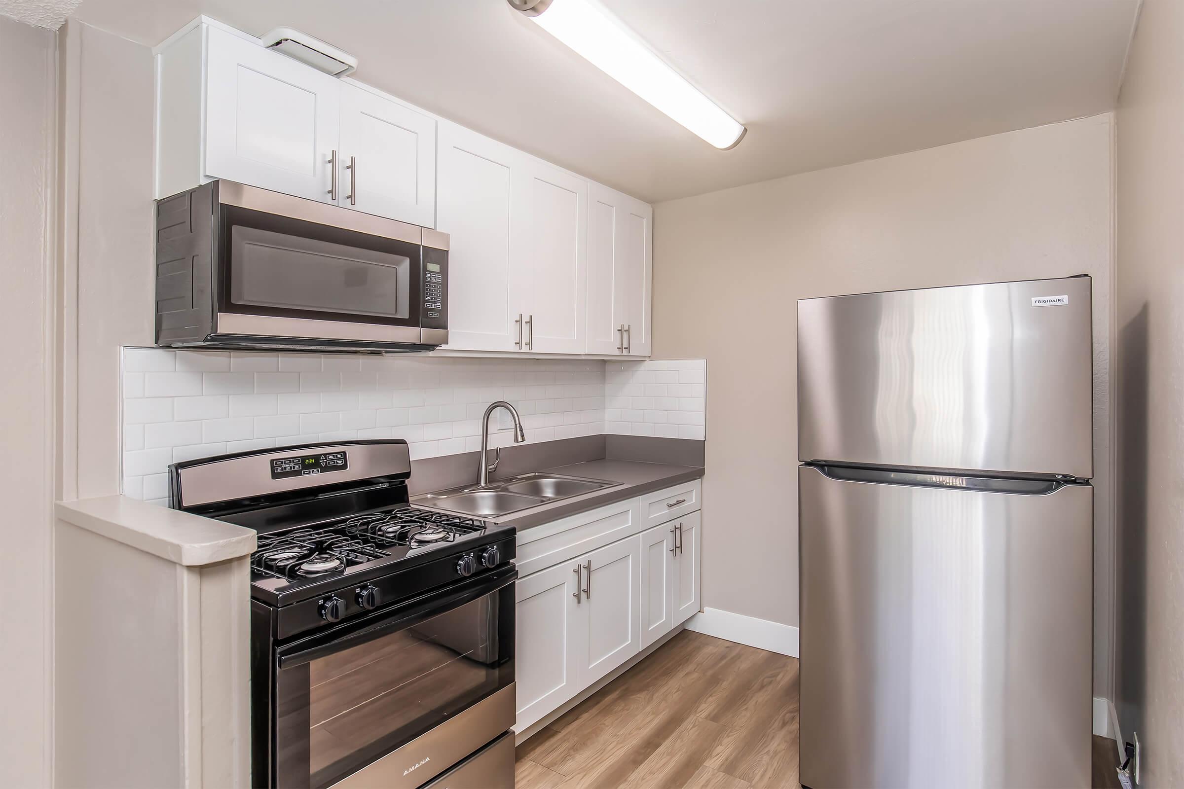 Modern kitchen with white cabinetry, black countertop, stainless steel appliances including a microwave, gas stove, and refrigerator. The kitchen features a sink with a sleek faucet and a neutral-colored wall. Ample lighting is provided by a ceiling fixture.