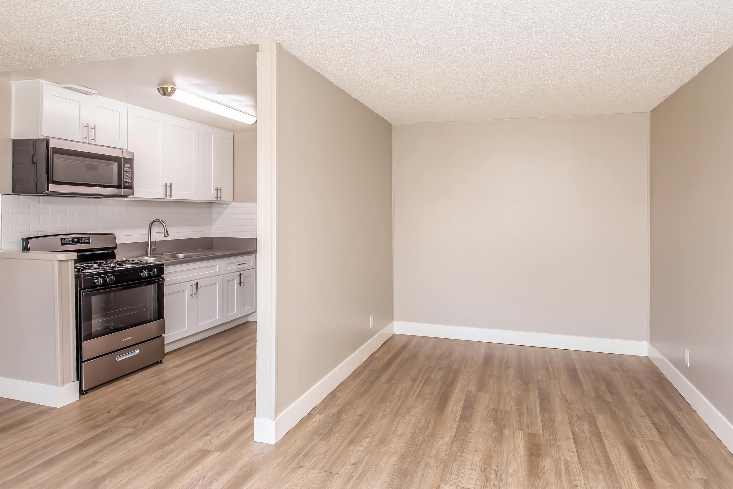 A modern kitchen with stainless steel appliances, including a gas stove and microwave, white cabinets, and a sink. Adjacent to the kitchen is a spacious living area with light brown wood flooring and neutral-colored walls, offering a bright and open feel.