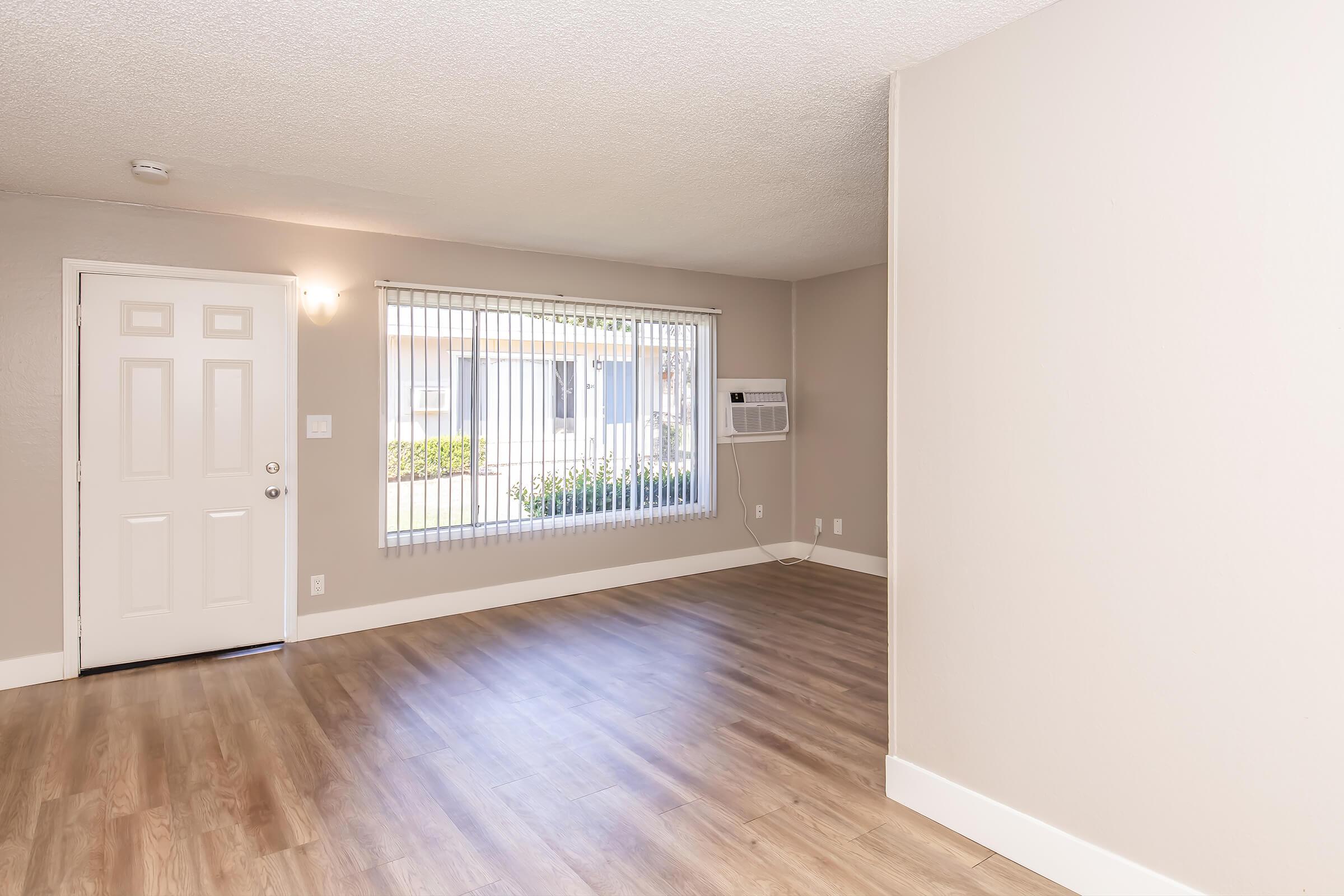A well-lit, empty living room with light-colored walls and wooden flooring. There is a door on the left leading outside, a large window with vertical blinds letting in natural light, and an air conditioning unit mounted below the window. The space is clean and ready for furnishings.