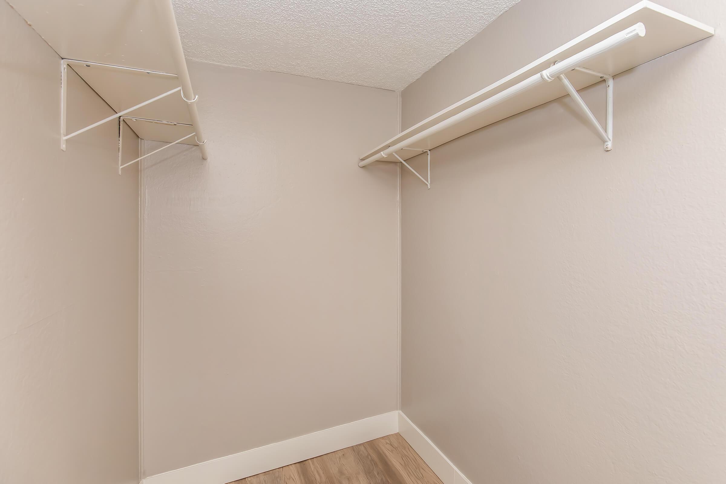 A minimalistic closet space with light beige walls, featuring two white wall-mounted shelves on opposite sides and a wooden floor.