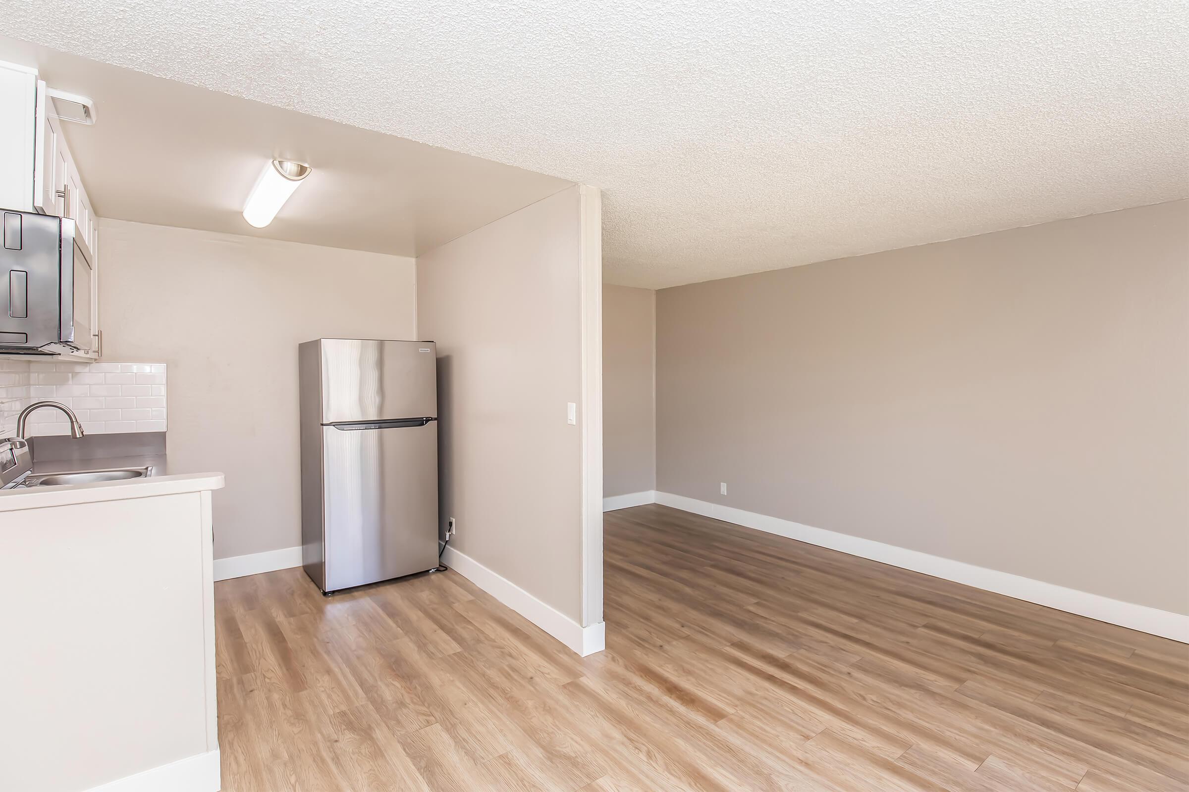 A spacious kitchen and living area featuring a stainless steel refrigerator, light-colored walls, and wood-like flooring. The kitchen includes a counter space with a sink, while the living area is open and unfurnished, creating a bright and airy atmosphere.