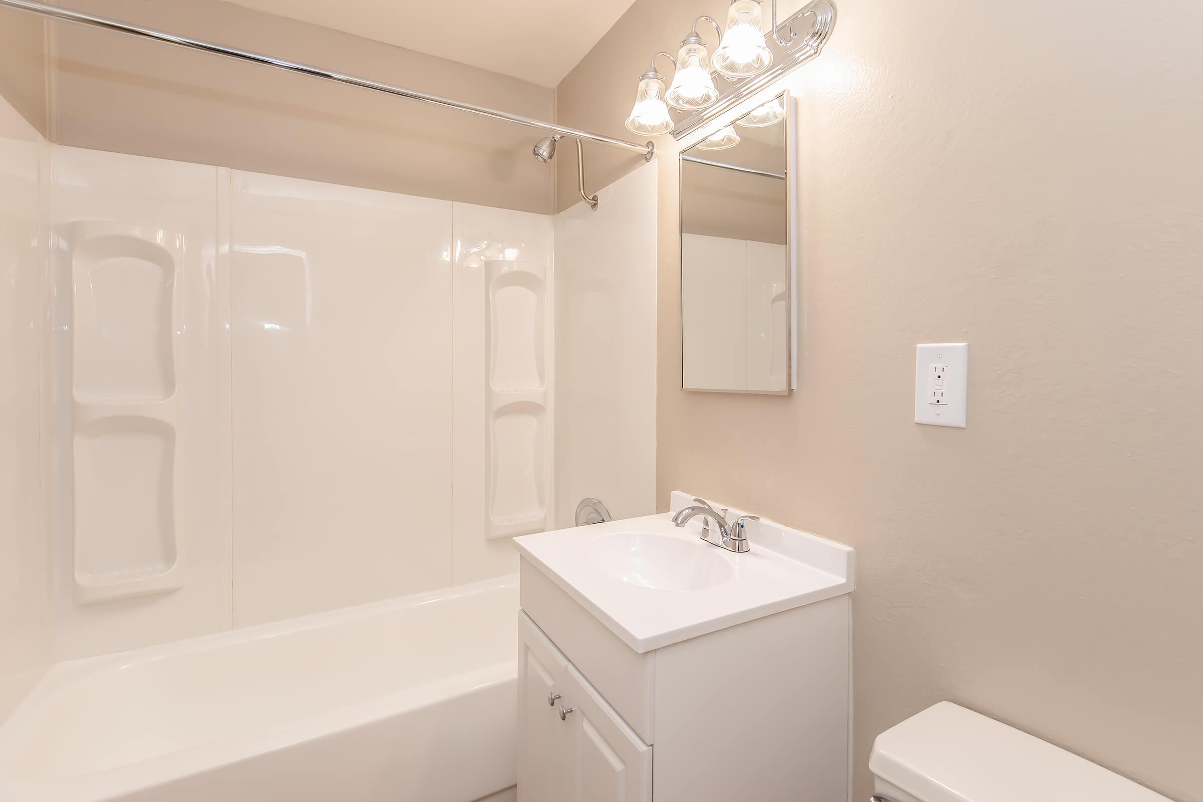 A clean, well-lit bathroom featuring a white bathtub with a shower, a small white vanity with a sink and mirror above, and a toilet. The walls are painted in a neutral color, and there are modern light fixtures above the mirror. The space appears tidy and inviting.