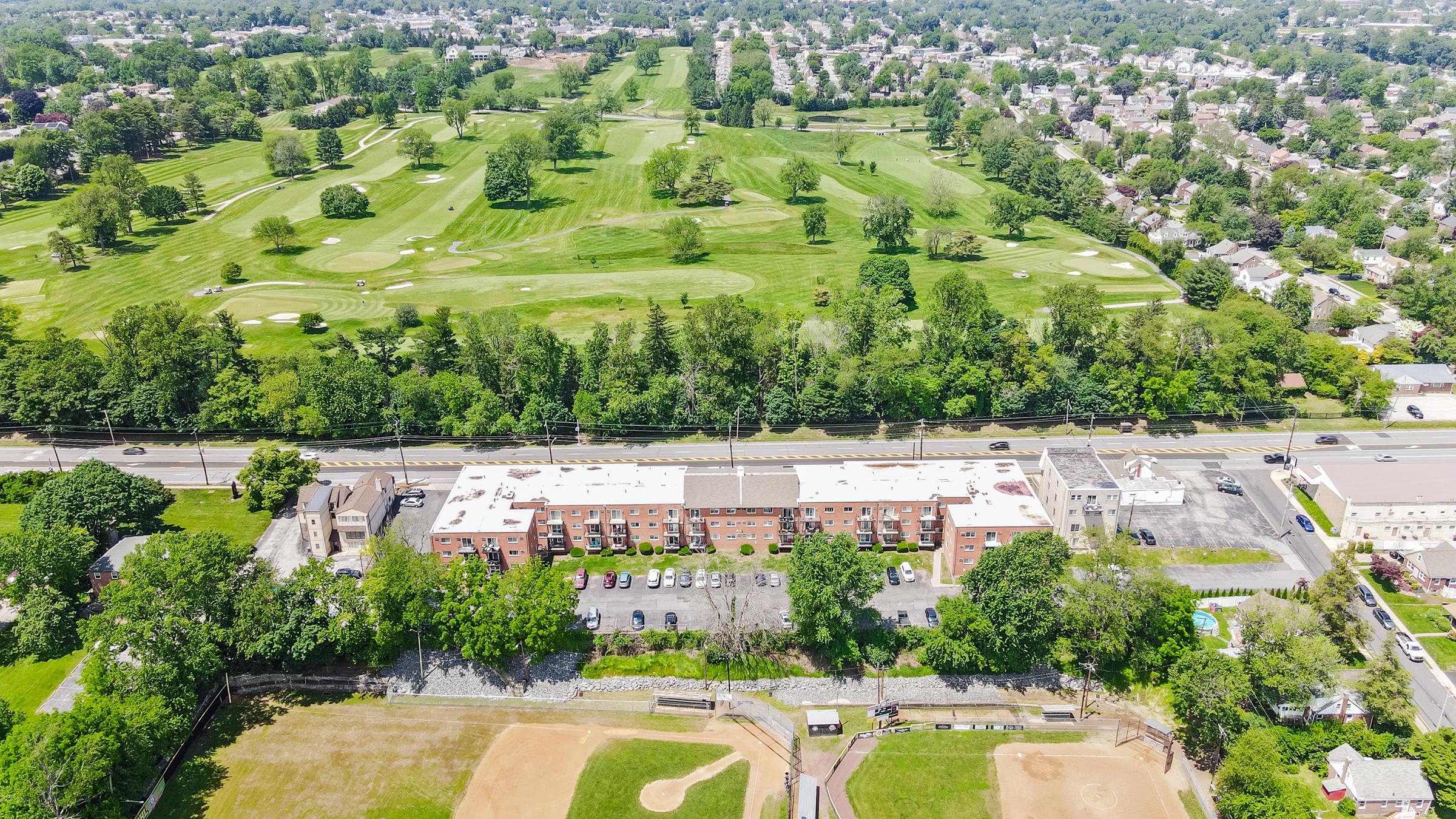 Aerial view of a large green golf course surrounded by trees, with a residential area in the background. In the foreground, there is a baseball field and an apartment complex with a parking lot. The scene captures a mix of recreational and residential spaces, showcasing lush greenery and urban development.