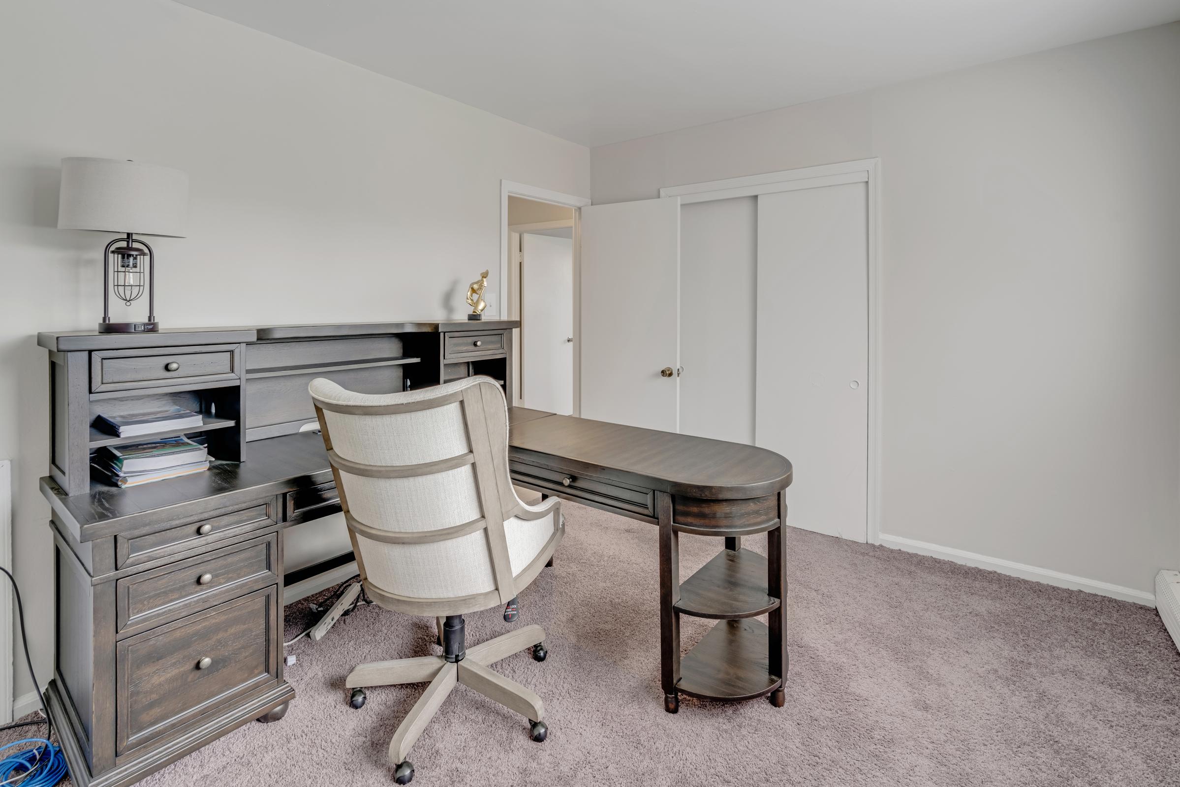 A minimalist home office with a dark wooden desk and a comfortable cream-colored swivel chair. The desk is equipped with a few items, and a lamp sits on the left. Behind the desk, a doorway leads to another room, while a plush carpet covers the floor, creating a cozy atmosphere.