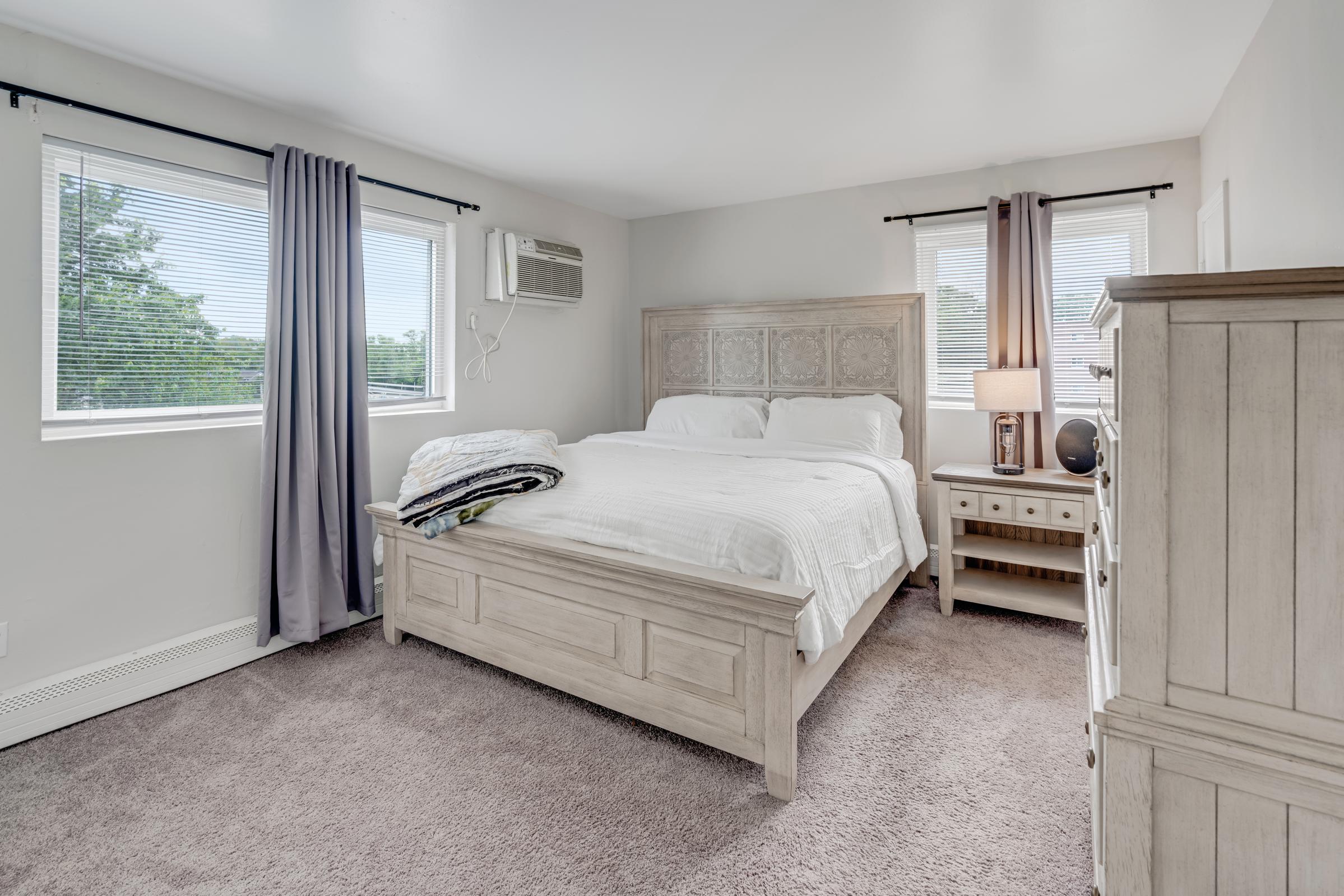 A cozy bedroom featuring a large bed with a light-colored headboard, soft white bedding, and bedside tables. Natural light streams in through two windows adorned with curtains. A dresser is positioned against one wall, and an air conditioning unit is visible. The carpeted floor adds warmth to the space.