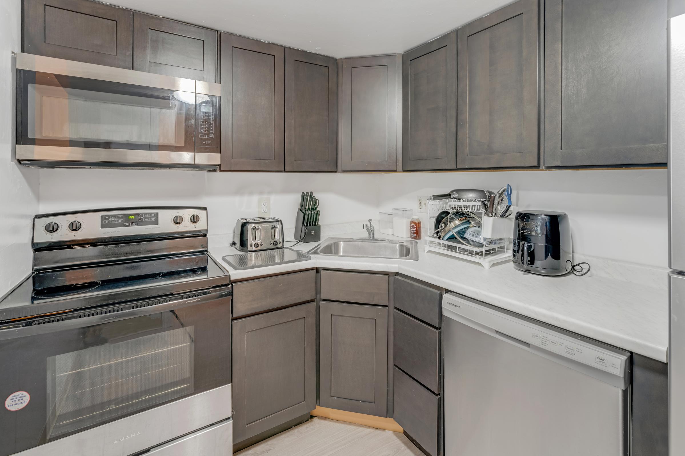 A modern kitchen featuring dark cabinets, stainless steel appliances including a microwave, oven, and dishwasher, a white sink, and a small toaster. The countertop is light-colored, and the kitchen is neatly arranged with dishware in a drying rack.