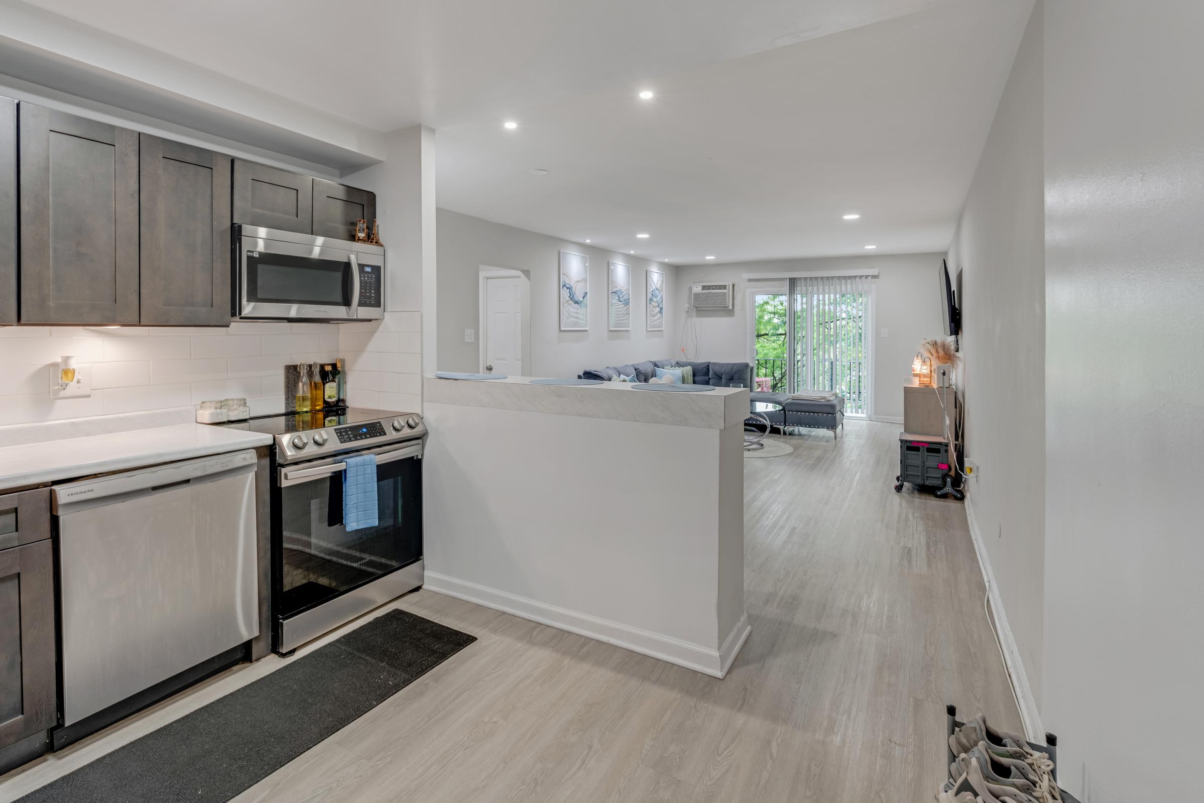 Modern kitchen with stainless steel appliances, including a stove, microwave, and dishwasher. Light wood cabinetry and a white tile backsplash. Open layout leads to a living area with a blue couch and natural light from sliding doors. Neutral color palette and contemporary decor throughout.