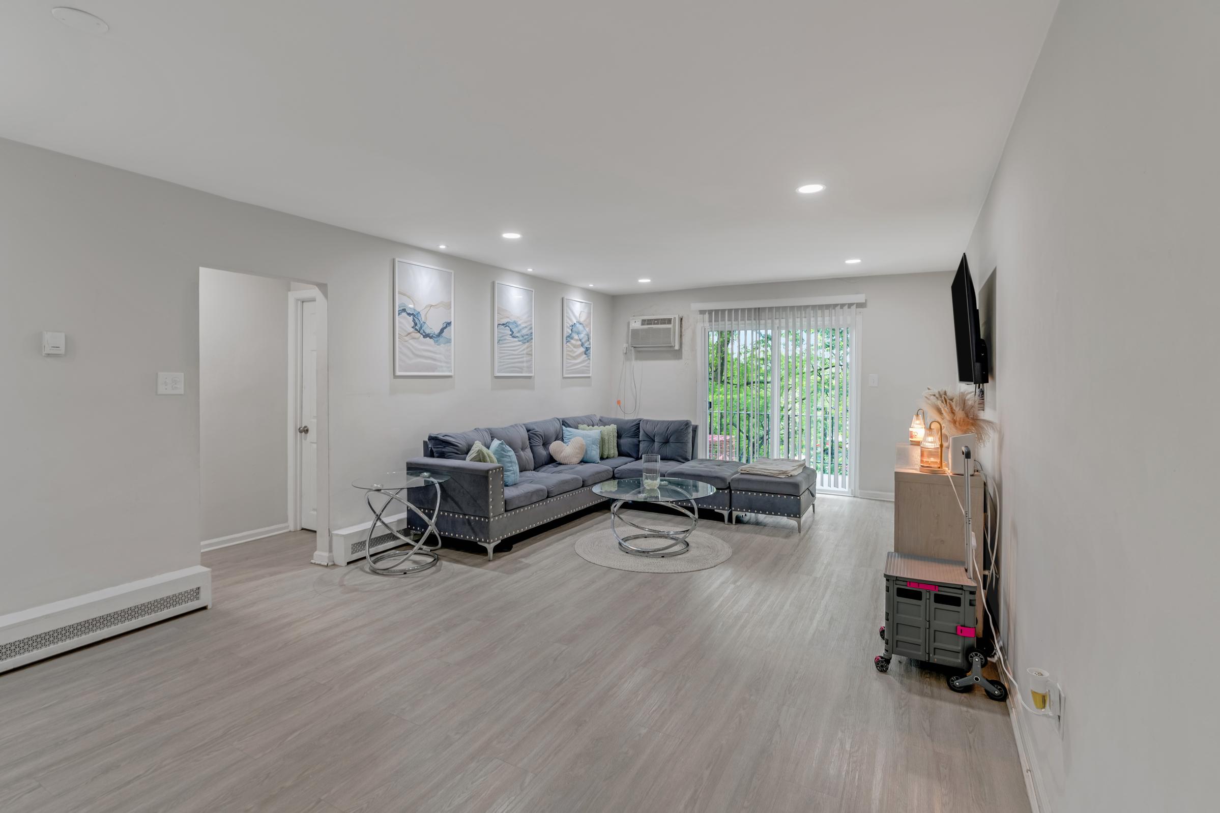 A modern, spacious living room featuring a gray sectional sofa, a glass coffee table, and artwork on the wall. There is a sliding glass door leading to an outdoor area, an air conditioning unit, and a television mounted on the wall, creating a bright and inviting atmosphere with light-colored walls and flooring.