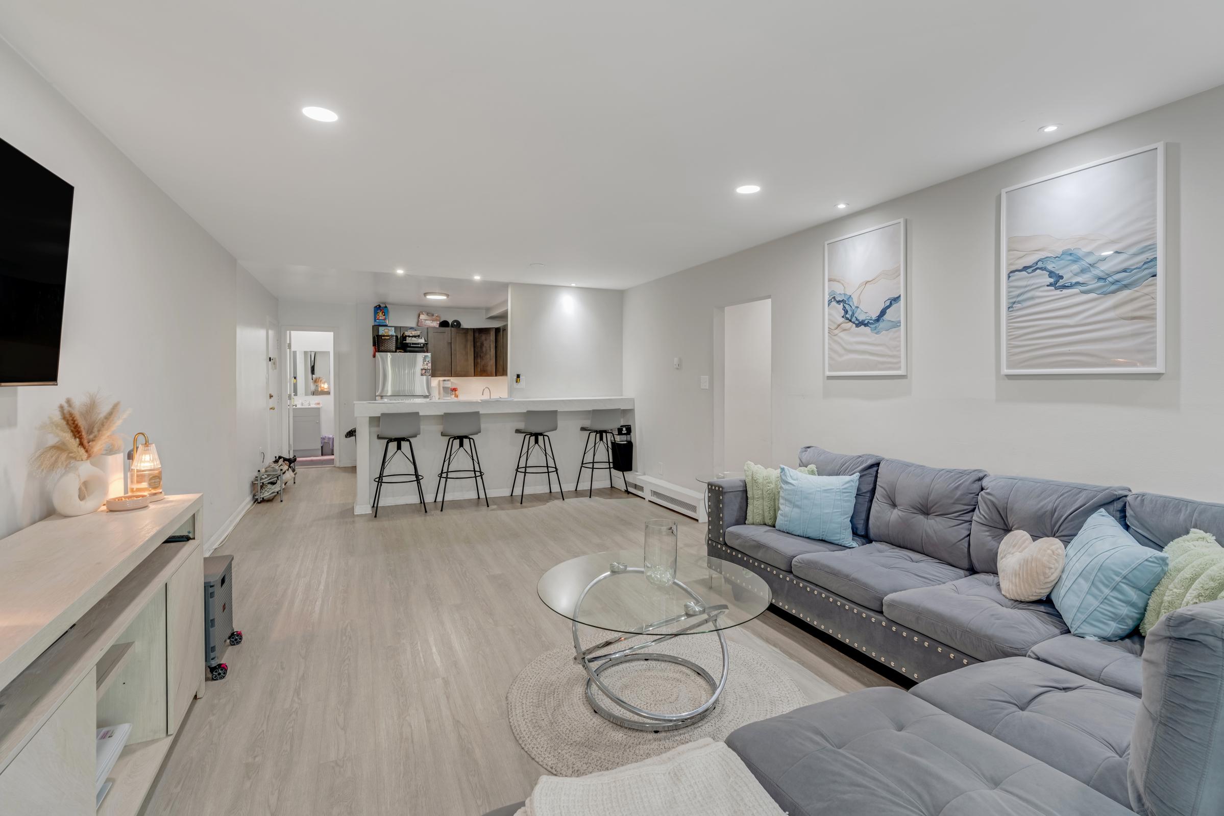 Modern living room featuring a gray sectional sofa with decorative pillows, a round glass coffee table, and a neutral area rug. The space is illuminated with soft lighting and has two large abstract wall art pieces. A view into a kitchen area with bar stools is visible in the background.