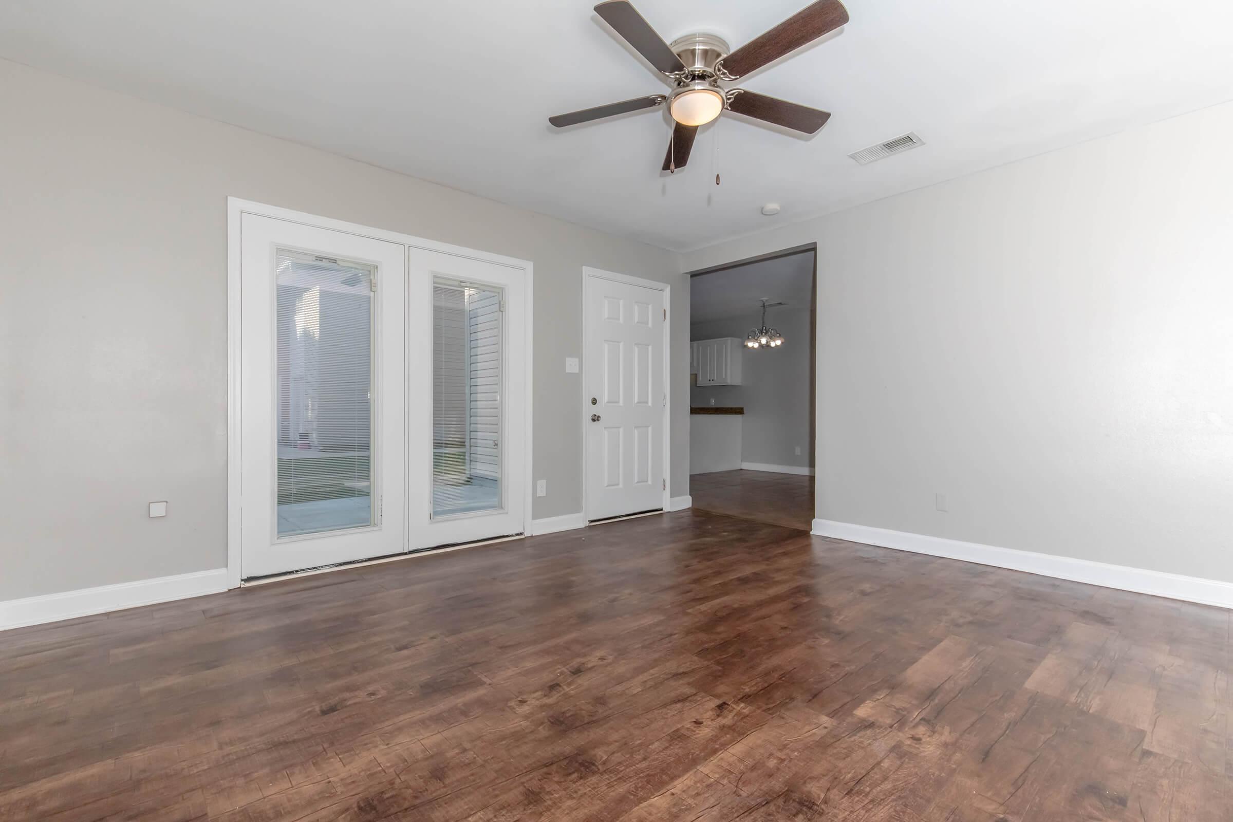 A spacious, light-colored living room featuring a wood floor, a ceiling fan, and large windows with French doors leading outside. The room has a neutral color palette, and an open doorway leads to another room, possibly a kitchen, which is visible in the background.