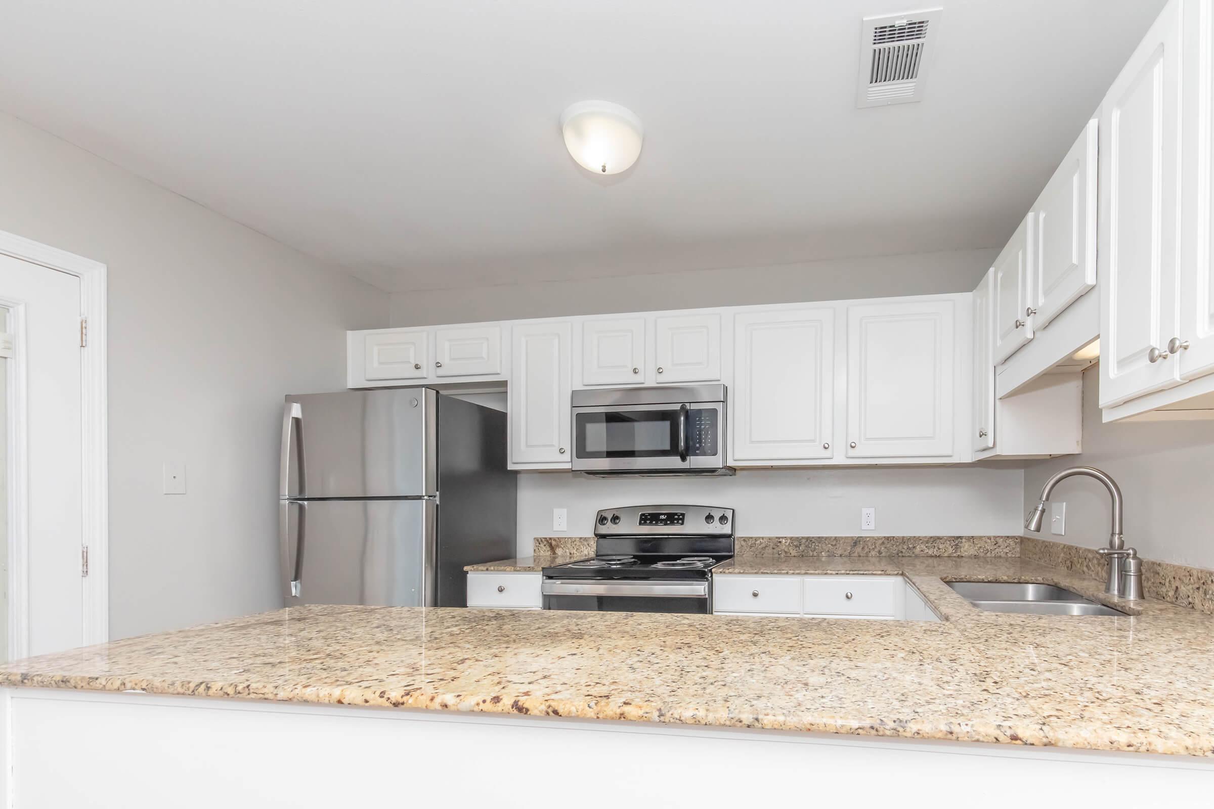 Modern kitchen featuring white cabinetry, granite countertops, stainless steel appliances including a refrigerator, microwave, and oven. The space is brightly lit with a ceiling light and has a sink under the window, providing a clean and inviting atmosphere.