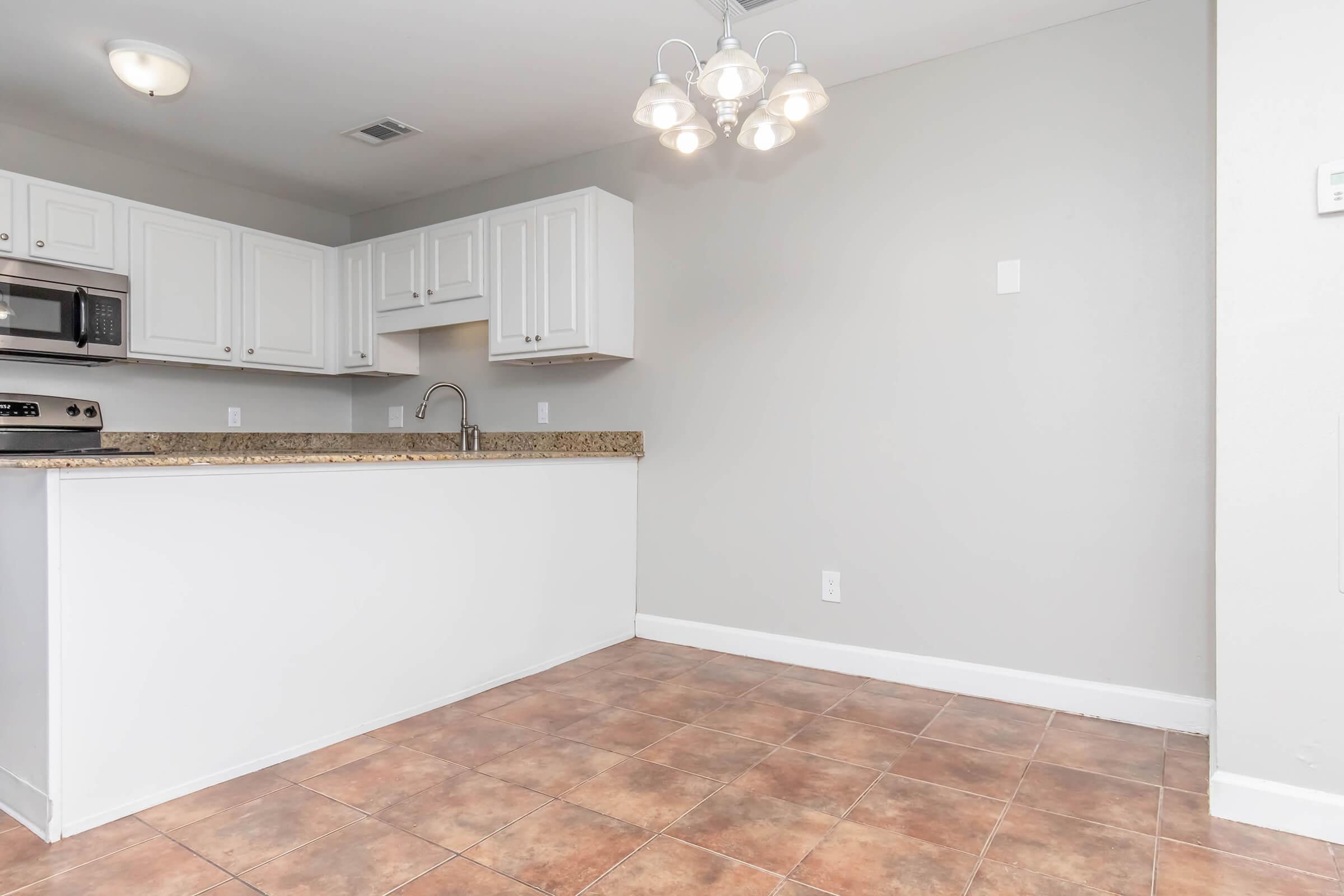 A modern kitchen featuring white cabinetry, a granite countertop, and stainless steel appliances. The space has beige tiled flooring and a ceiling light fixture with several bulbs. The walls are painted in a light gray color, creating a clean and contemporary atmosphere.