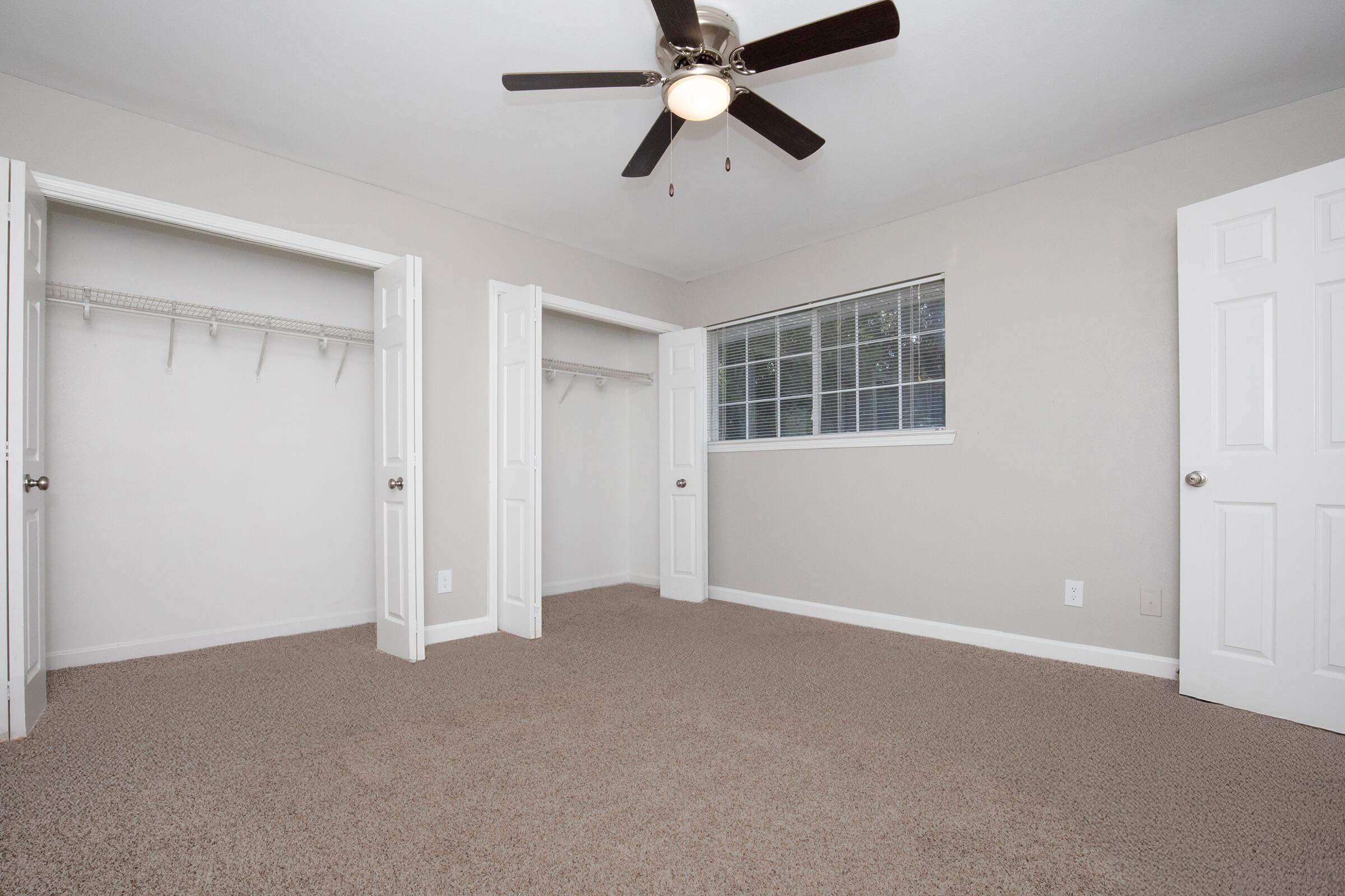 A spacious bedroom with light gray walls, a ceiling fan, and plush carpet. Two white doors lead to empty closets on either side, and a window brings in natural light. The room has a clean and airy feel, perfect for personalization.