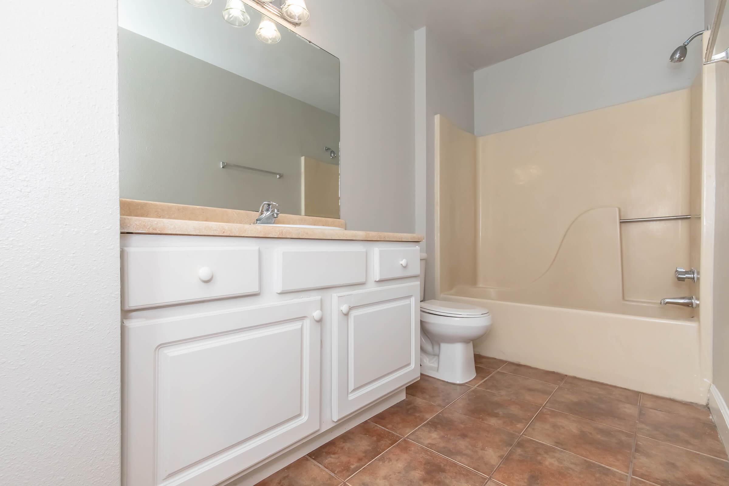 A clean and simple bathroom featuring a white vanity with a sink, a large mirror above it, a light-colored bathtub with a showerhead, and a white toilet. The floor is tiled in a brown pattern, and the walls are painted in neutral tones, creating a spacious and bright appearance.