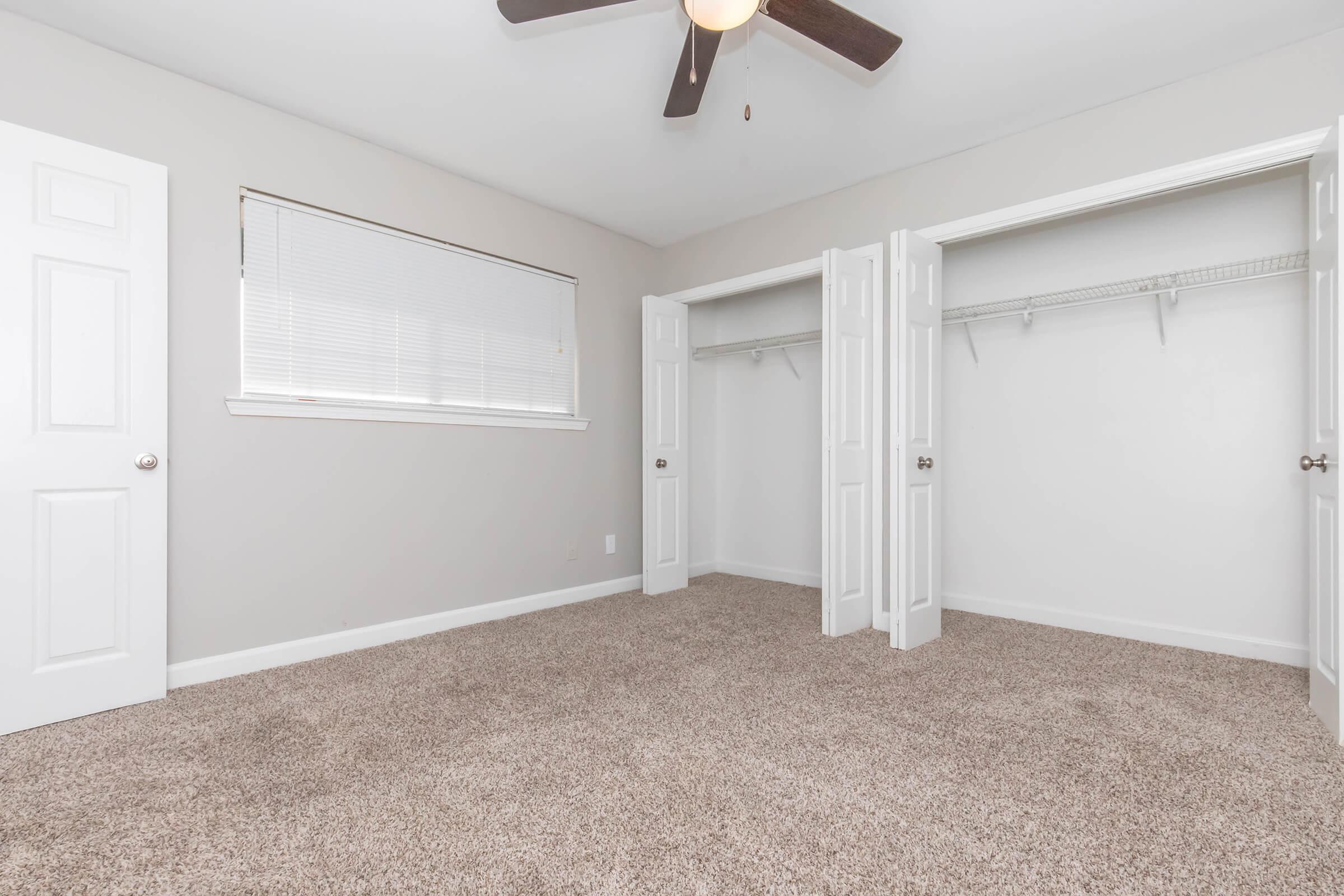 Empty bedroom featuring beige walls, plush carpet, a ceiling fan with wooden blades, and two open closets with white shelving. A window with blinds allows natural light to enter the space.