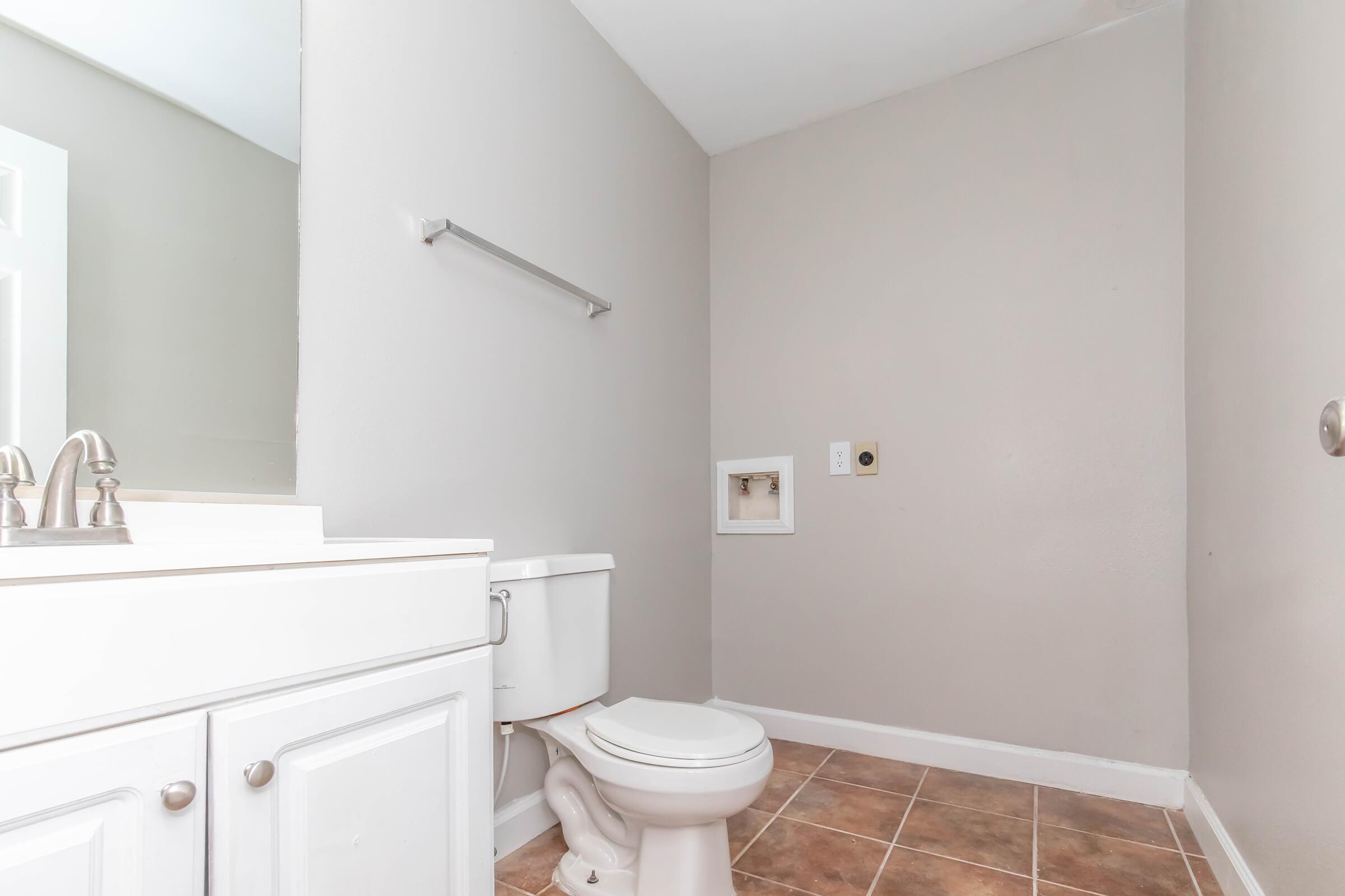 A clean bathroom featuring a white toilet, a white sink with cabinets, and beige tiled flooring. The walls are painted in a light gray color, and there is a mirror above the sink. A towel bar is mounted on the wall, and an electrical outlet is visible near the floor. The room appears well-lit and uncluttered.