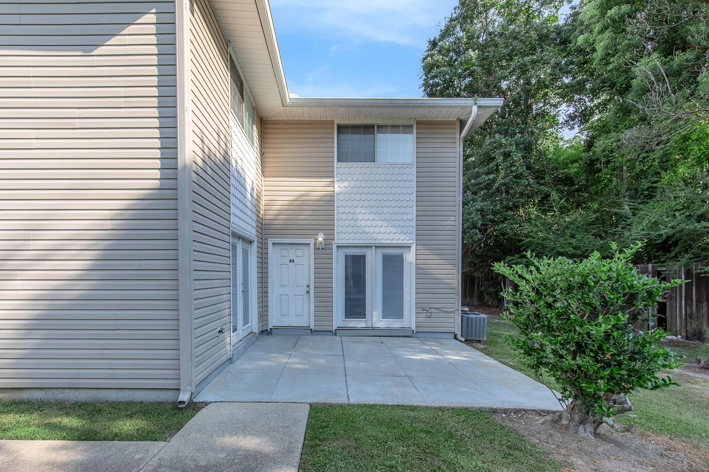 A view of the exterior of a two-story building featuring a light-colored siding, a patio area with concrete flooring, and two doors leading into the building. There is a small shrub in the foreground and trees in the background, creating a green, natural environment.