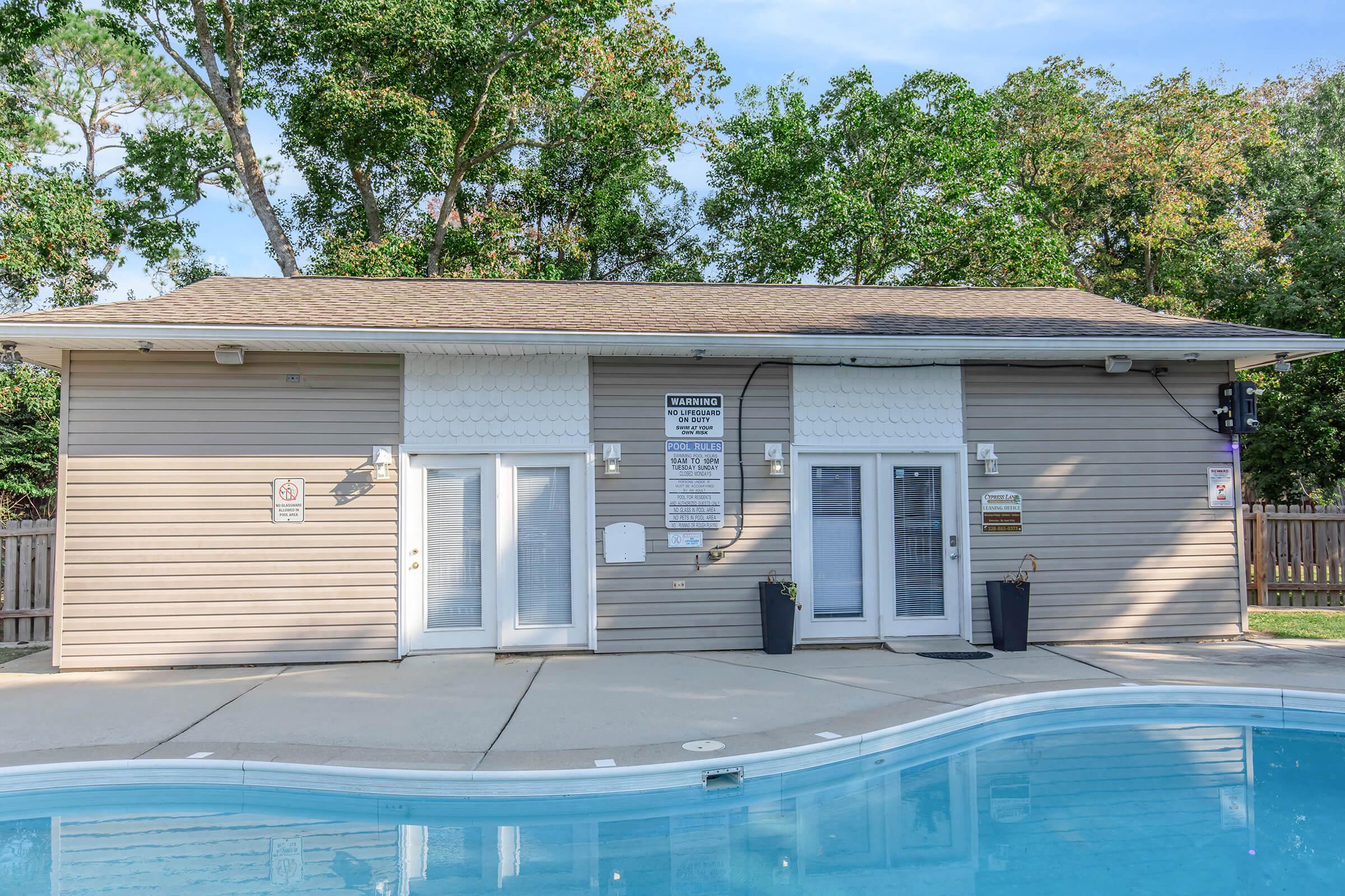 A swimming pool area featuring a small building with a sign. The building has double doors and windows, surrounded by trees and greenery. The pool is clear with a concrete area, and planters are positioned near the entrance.