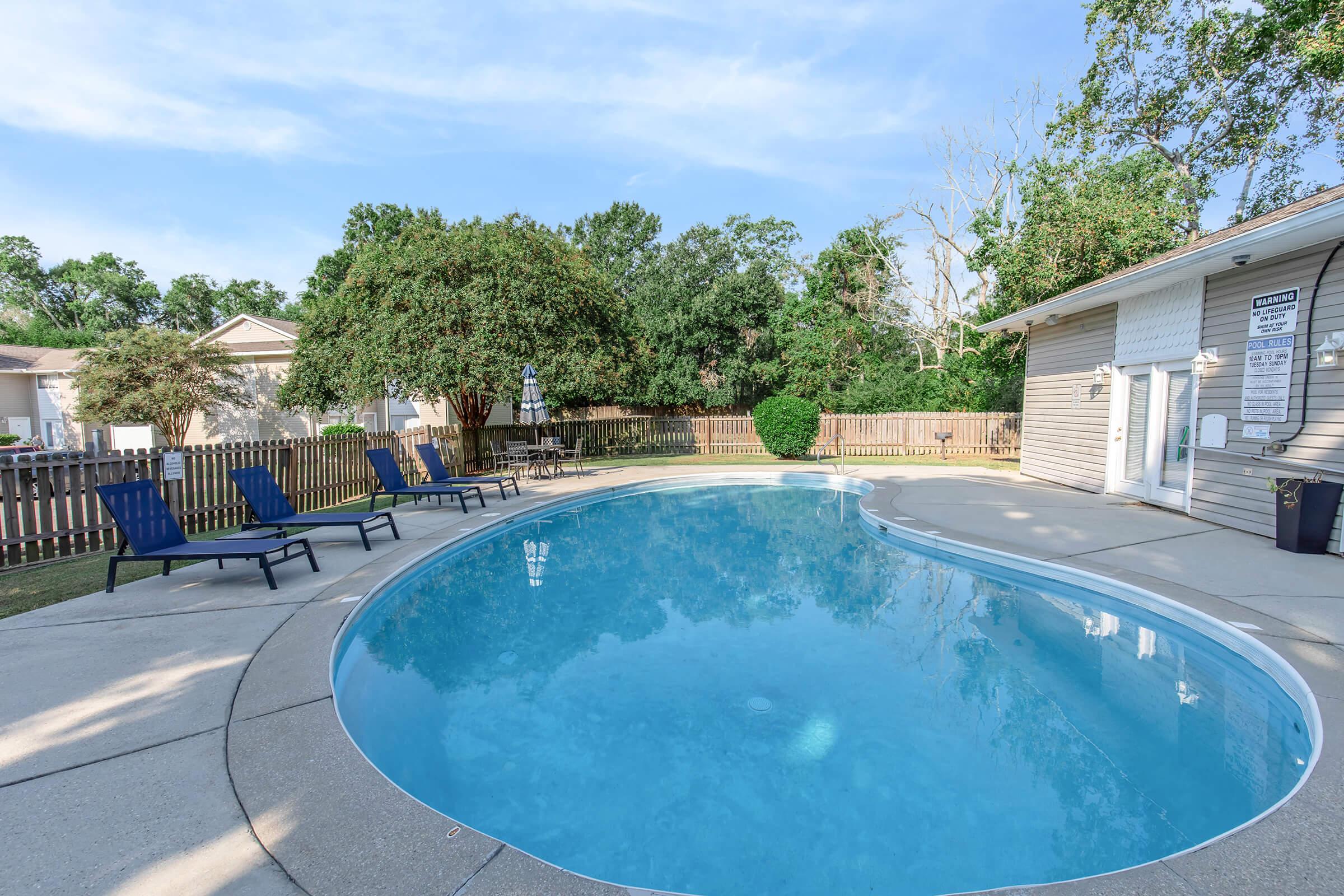 A clean, inviting swimming pool surrounded by lounge chairs, set in a well-maintained outdoor area. Lush greenery and a wooden fence provide privacy. The sky is clear, enhancing the tranquil atmosphere of the poolside space. Perfect for relaxation and leisure.