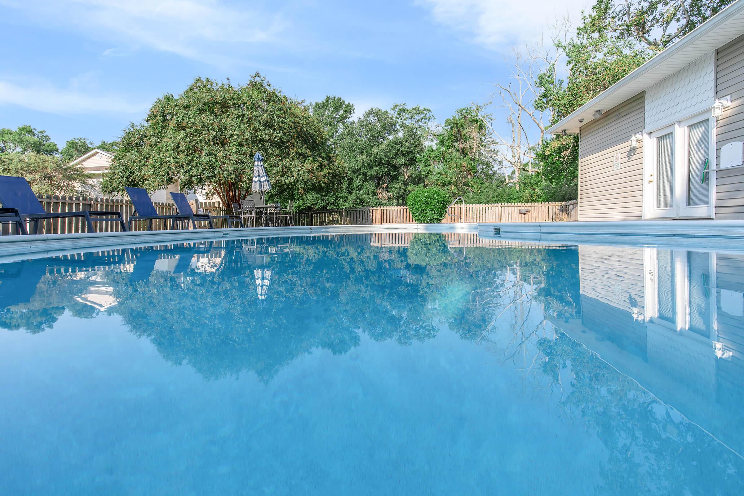 A serene swimming pool with clear blue water reflecting the sky, surrounded by lush greenery and a wooden fence. Sun loungers are positioned nearby, and a house with light-colored siding is visible in the background. The scene conveys a tranquil outdoor setting ideal for relaxation.