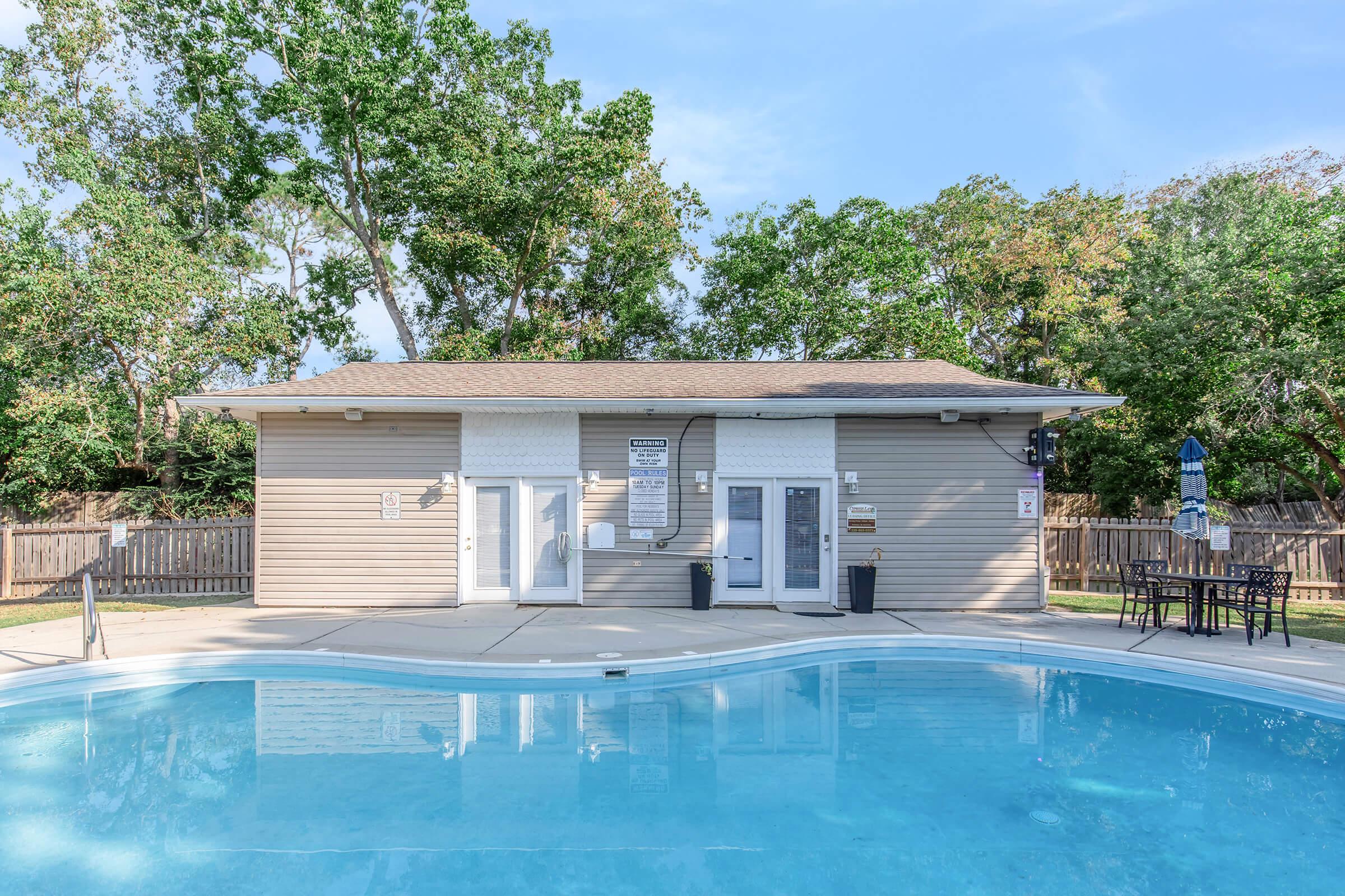 A clear blue swimming pool in front of a modern, single-story building with light-colored siding. The building features large windows and doors, and is surrounded by green trees and a wooden fence. Outdoor seating with tables is near the pool area, creating a tranquil atmosphere for relaxation.