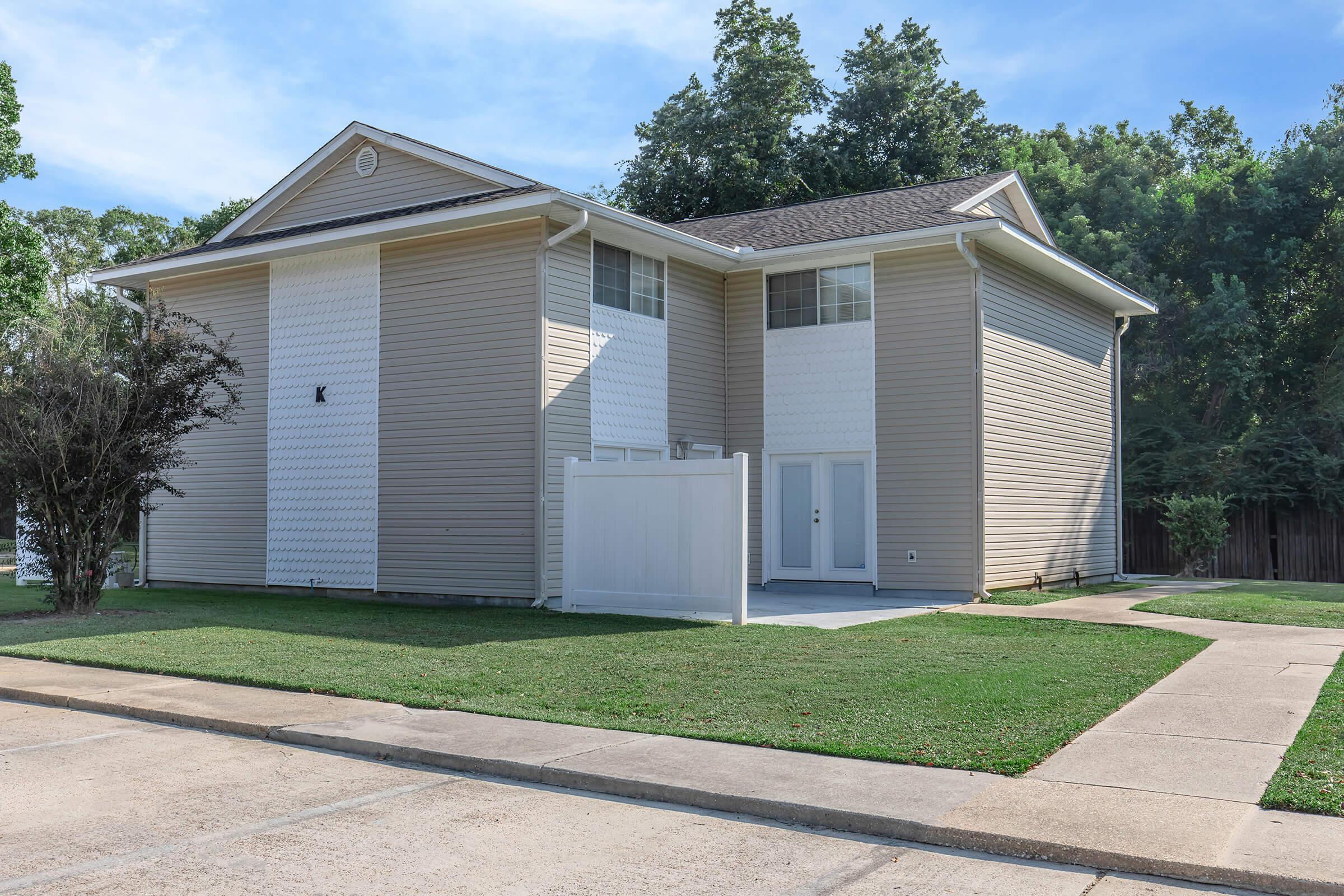 Two-story residential house with light beige siding, white accents, and a fenced yard. The front features a walkway leading to the entrance, flanked by green grass, and is set against a backdrop of trees. Blue sky with scattered clouds overhead.