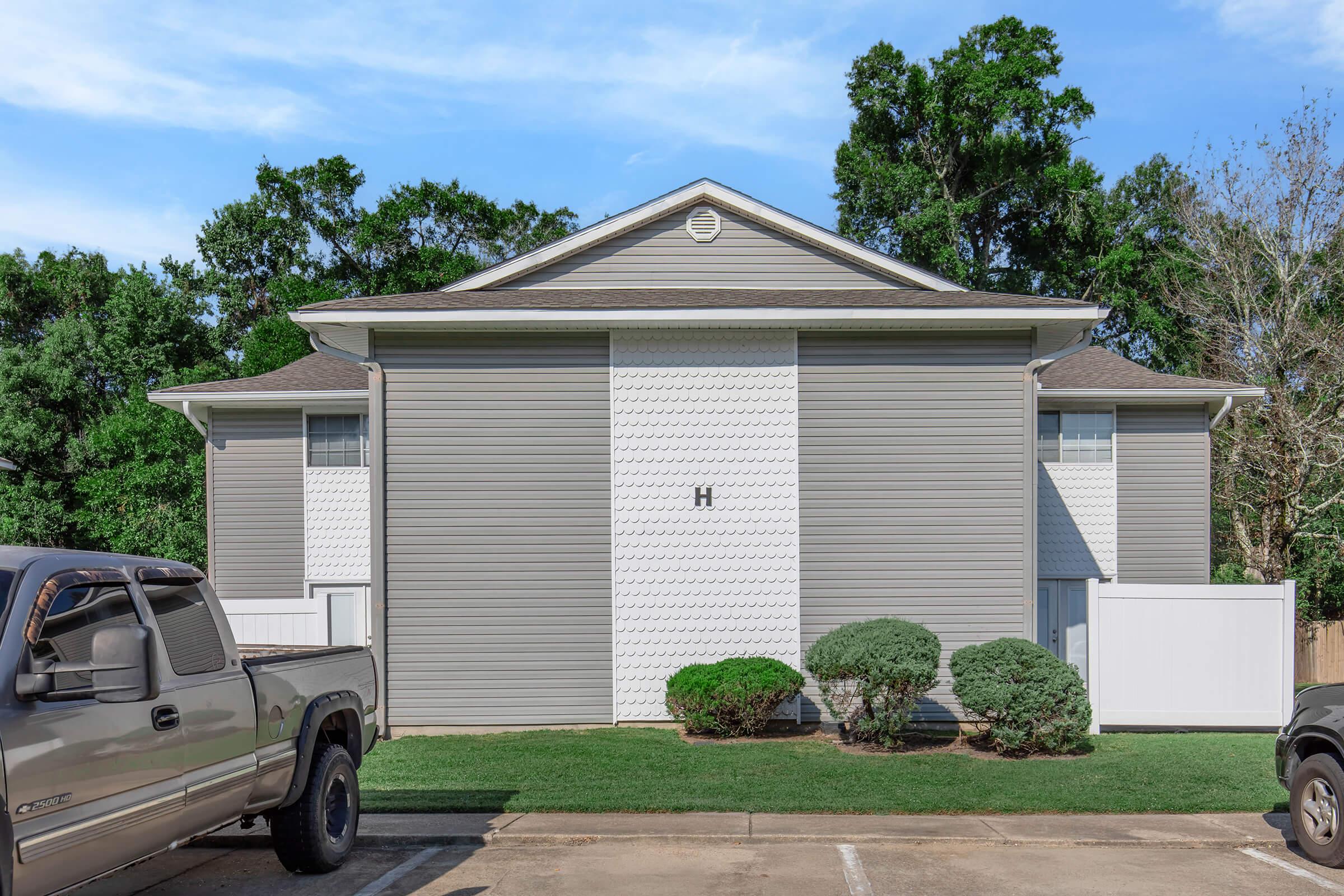 A two-story residential building with a light gray exterior, featuring a symmetrical design. The front yard is grassy with two small bushes, and there are parked vehicles in the foreground. The backdrop includes trees and a clear blue sky.
