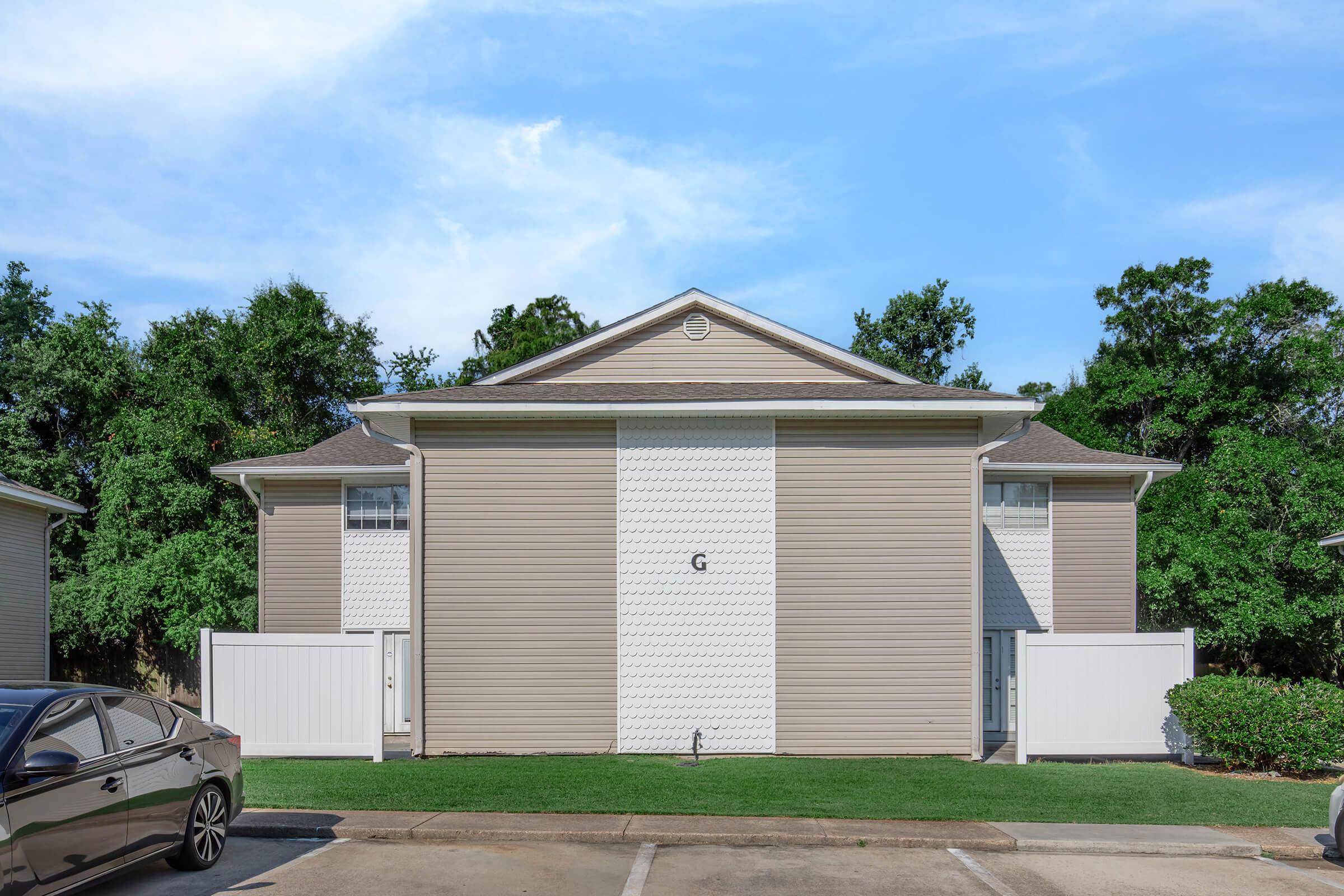 Two-story residential building with beige siding, featuring a central white panel and a letter "G" displayed. The property is flanked by white fences and surrounded by green trees. A parked black car is visible in the foreground, and the sky is clear with some clouds.
